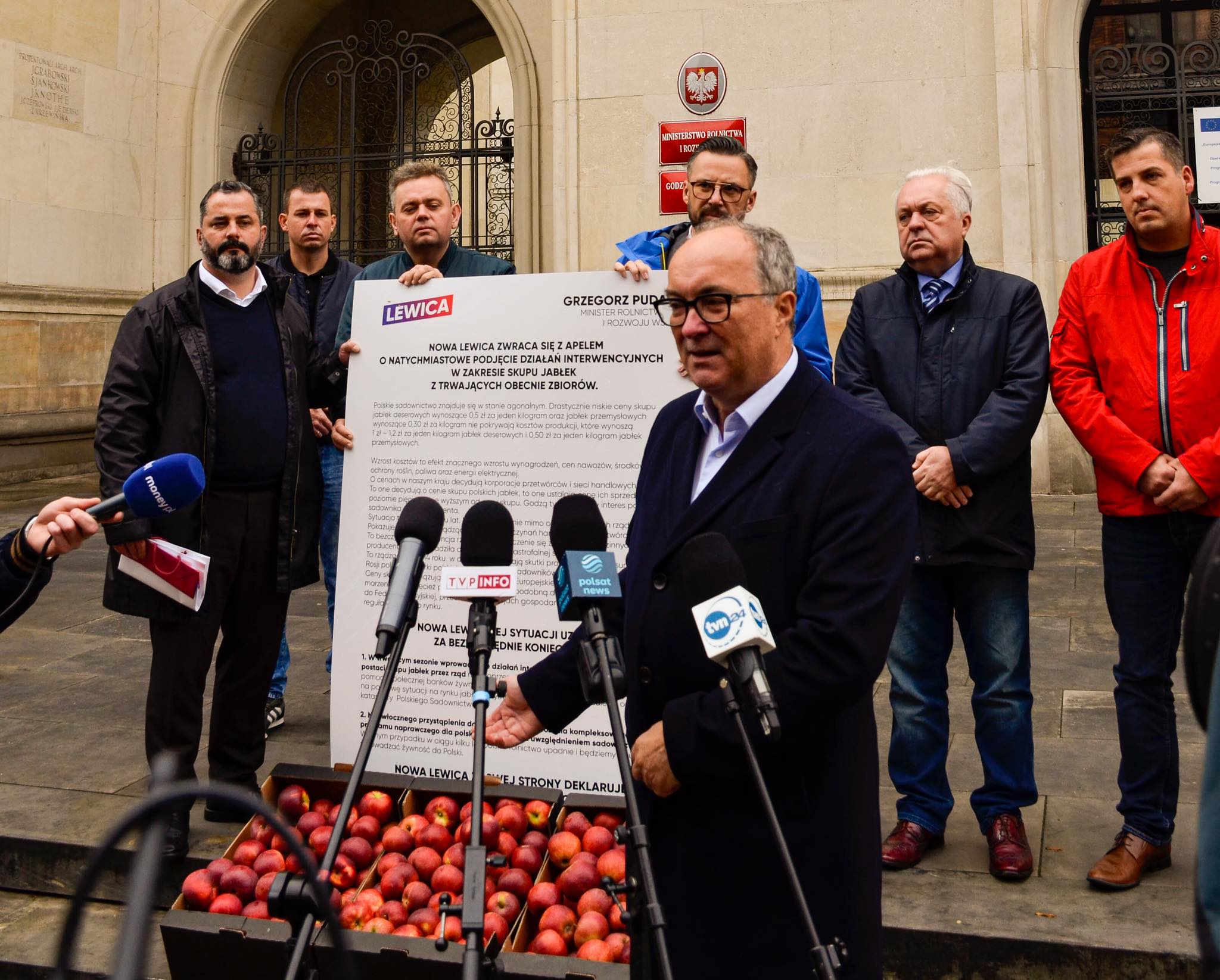 Grupa osób stojących na konferencji prasowej Lewicy przed budynkiem, wokół której ustawiono mikrofony i duży plakat o interwencyjnym skupie jabłek, z widocznymi skrzynkami pełnymi jabłek na pierwszym planie.