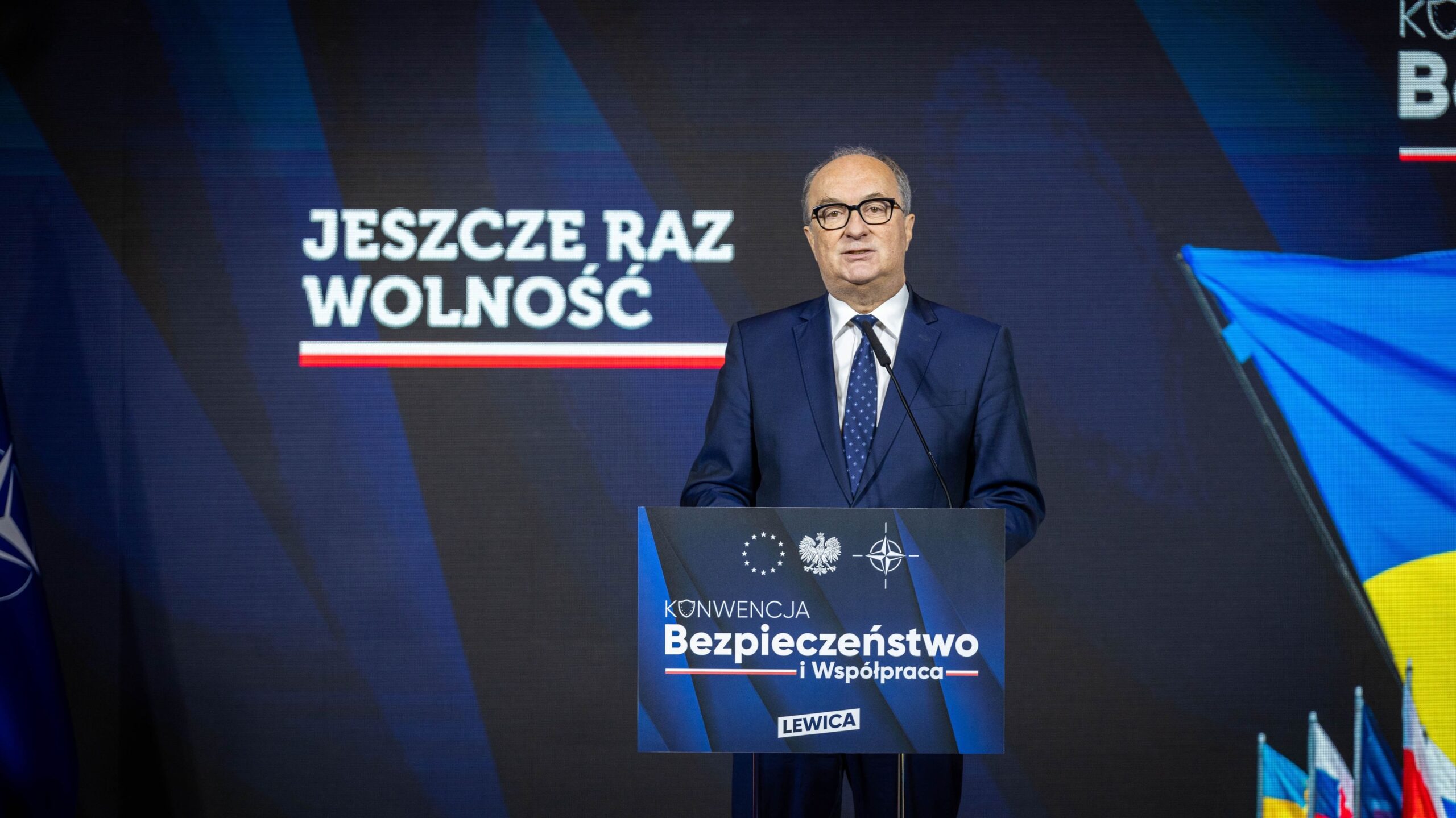 Mężczyzna w granatowym garniturze przemawia z mównicy podczas konferencji o kompleksowym planie wzmocnienia bezpieczeństwa Polski, na tle ekranu z hasłem „Jeszcze raz wolność” i motywem „Bezpieczeństwo i Współpraca”.