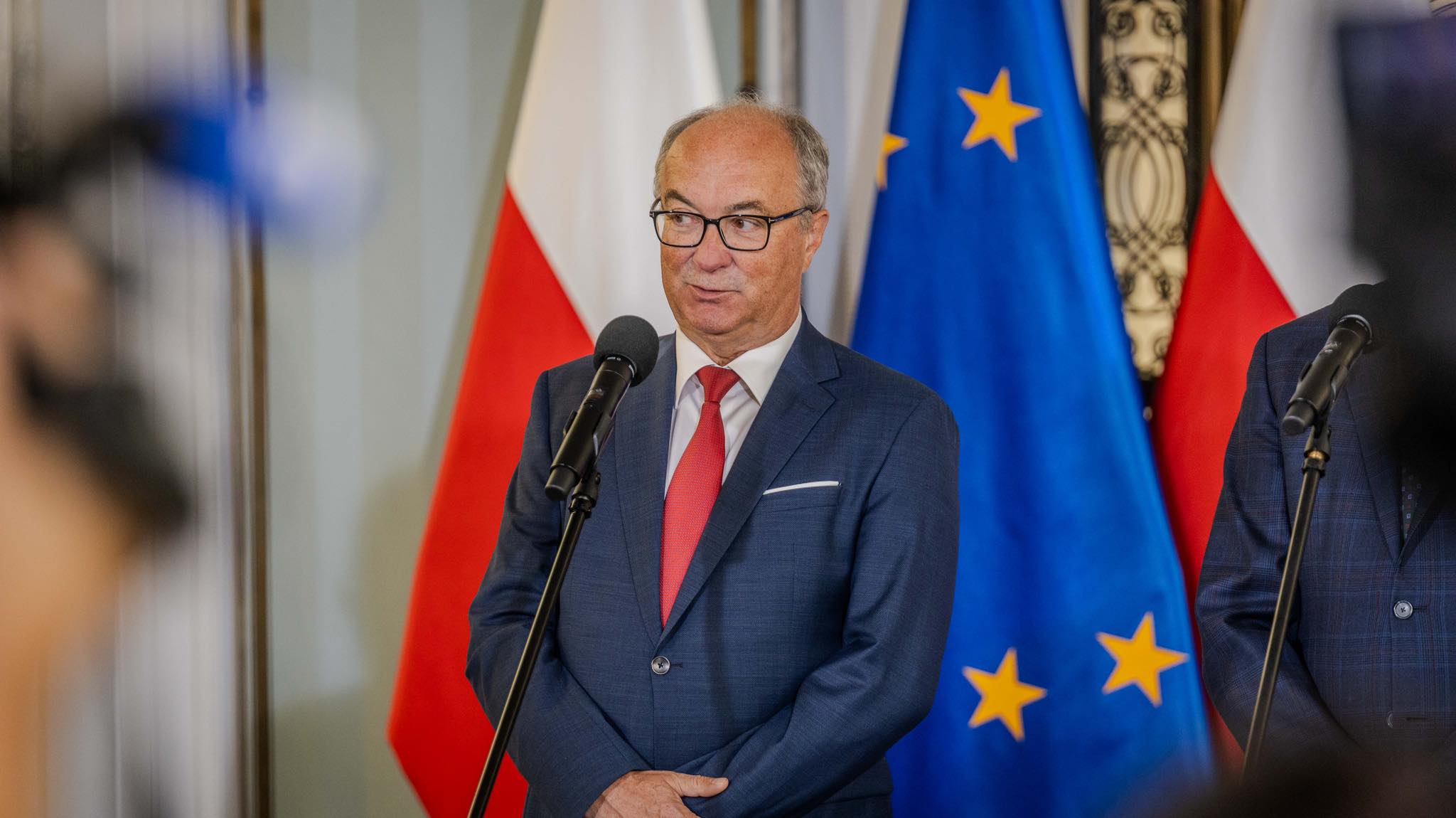 Polityk opozycji w granatowym garniturze i czerwonym krawacie przemawia do mikrofonów podczas konferencji prasowej, w tle widoczne flagi Polski i Unii Europejskiej.