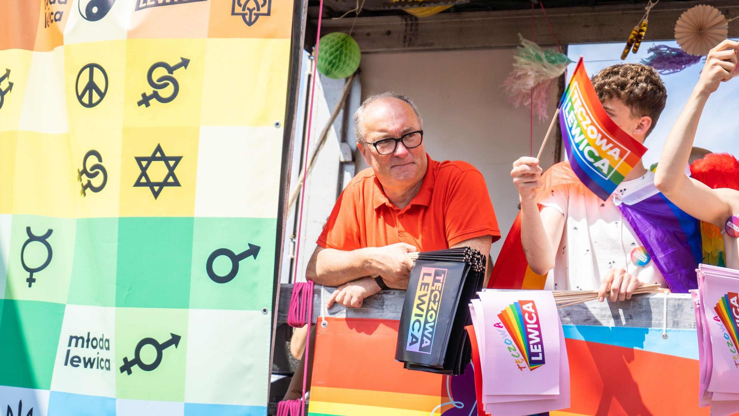 Mężczyzna w pomarańczowej koszulce stoi przy barierce, trzymając ulotki i obserwując wydarzenie związane z ruchem LGBTQ+, otoczony kolorowymi flagami i banerami.