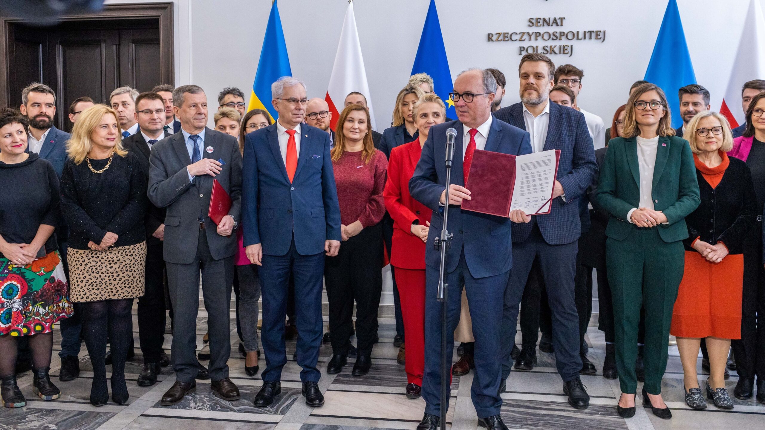 Grupa polityków Lewicy stoi razem podczas podpisywania koalicyjnego porozumienia między Nową Lewicą, Razem, PPS i Unią Pracy, na tle flag państwowych.