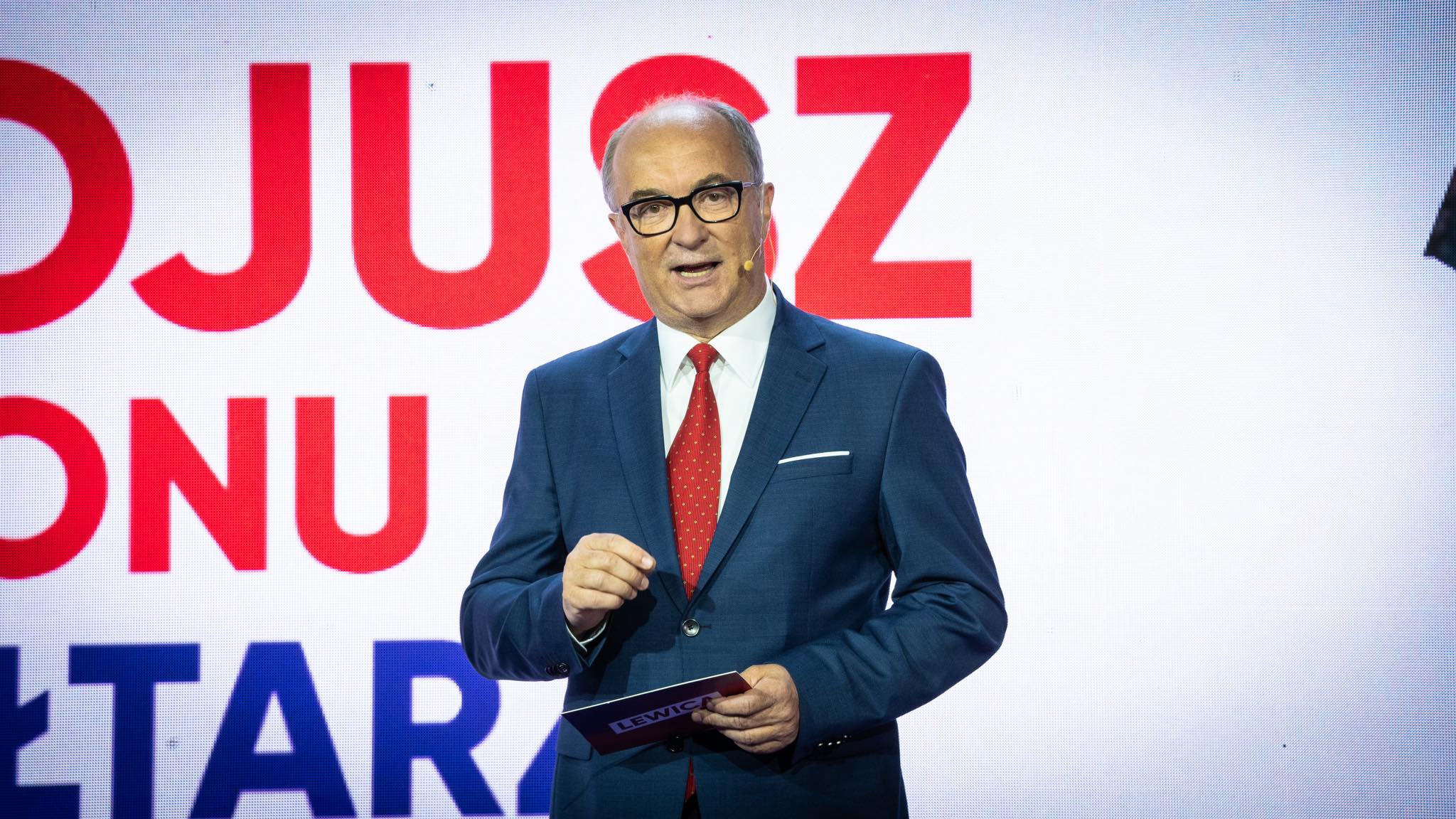 Mężczyzna w granatowym garniturze, białej koszuli i czerwonym krawacie przemawia na scenie, trzymając kartkę z napisem Lewica, na tle dużych czerwonych liter.