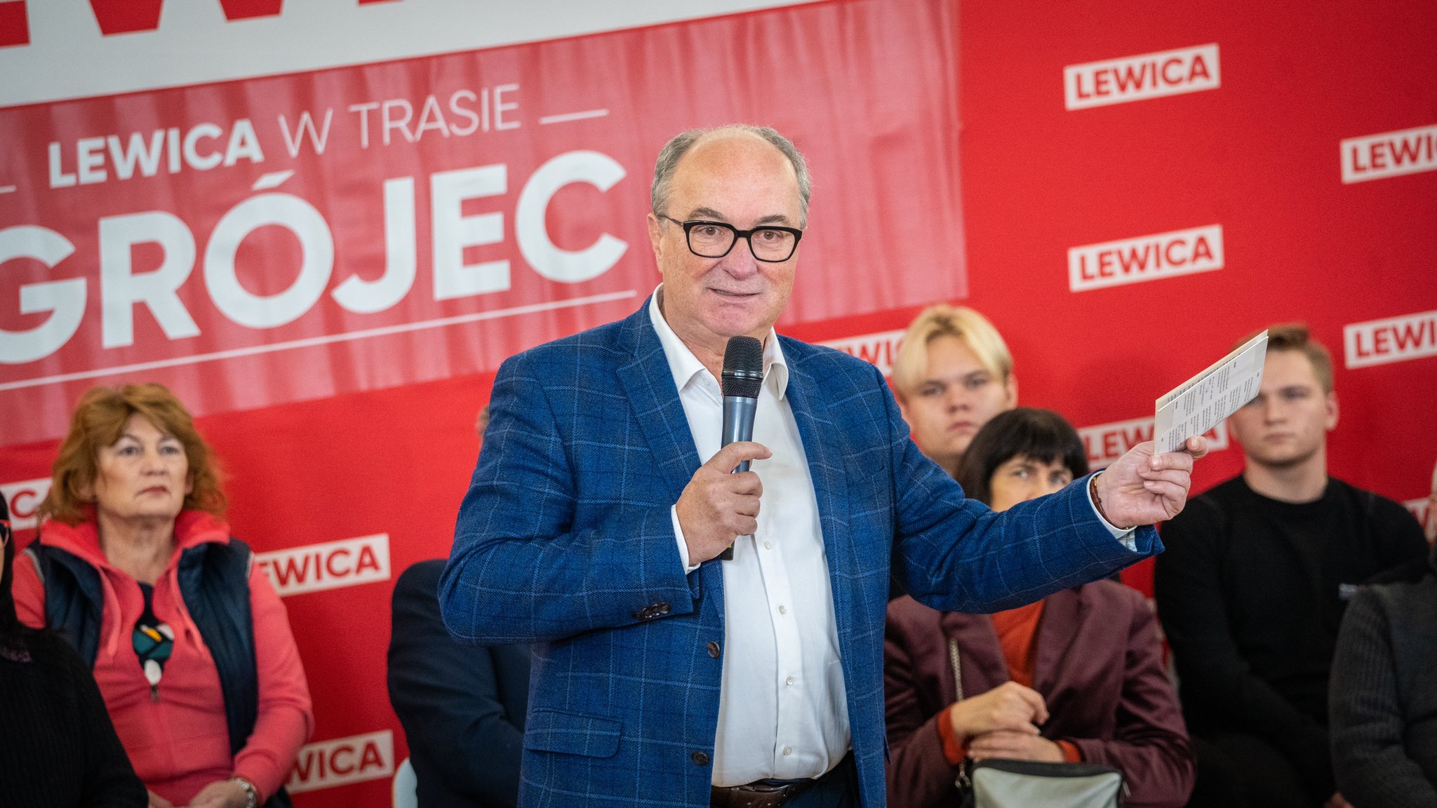 Na zdjęciu widać mężczyznę w okularach, który przemawia do publiczności podczas wydarzenia zorganizowanego przez Lewicę, a w tle widoczny jest czerwony baner z napisem 