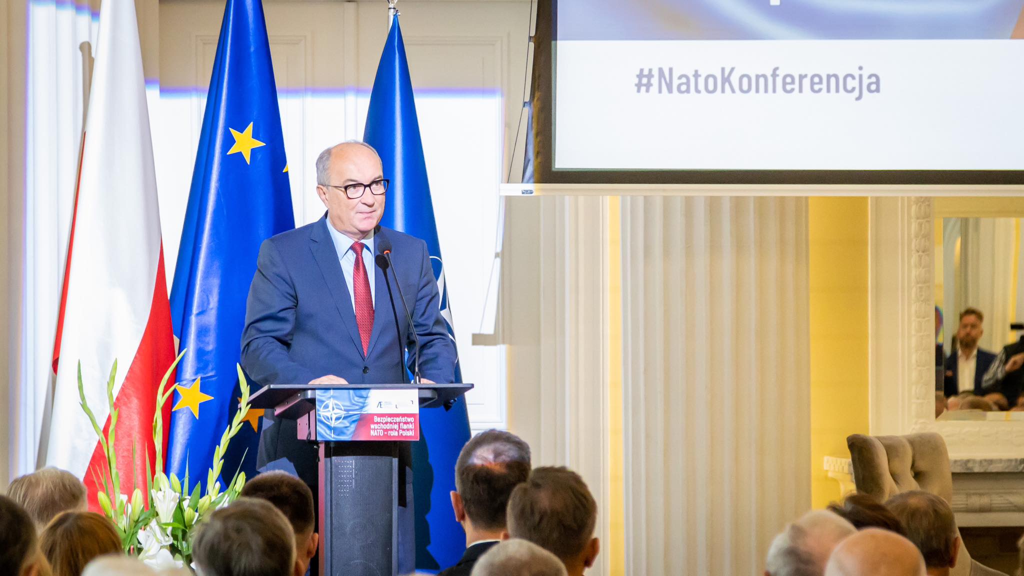 Na zdjęciu widać mężczyznę przemawiającego podczas konferencji dotyczącej NATO, z flagami Polski i Unii Europejskiej w tle.