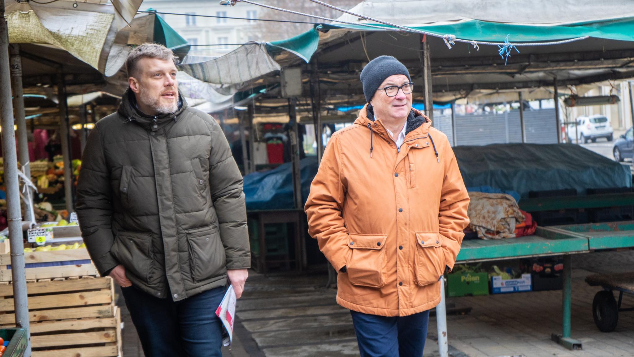 Dwóch mężczyzn spaceruje po targu w Wielkopolsce, jeden w ciemnozielonej kurtce, drugi w pomarańczowej kurtce, w tle stoją stoiska z owocami i warzywami.