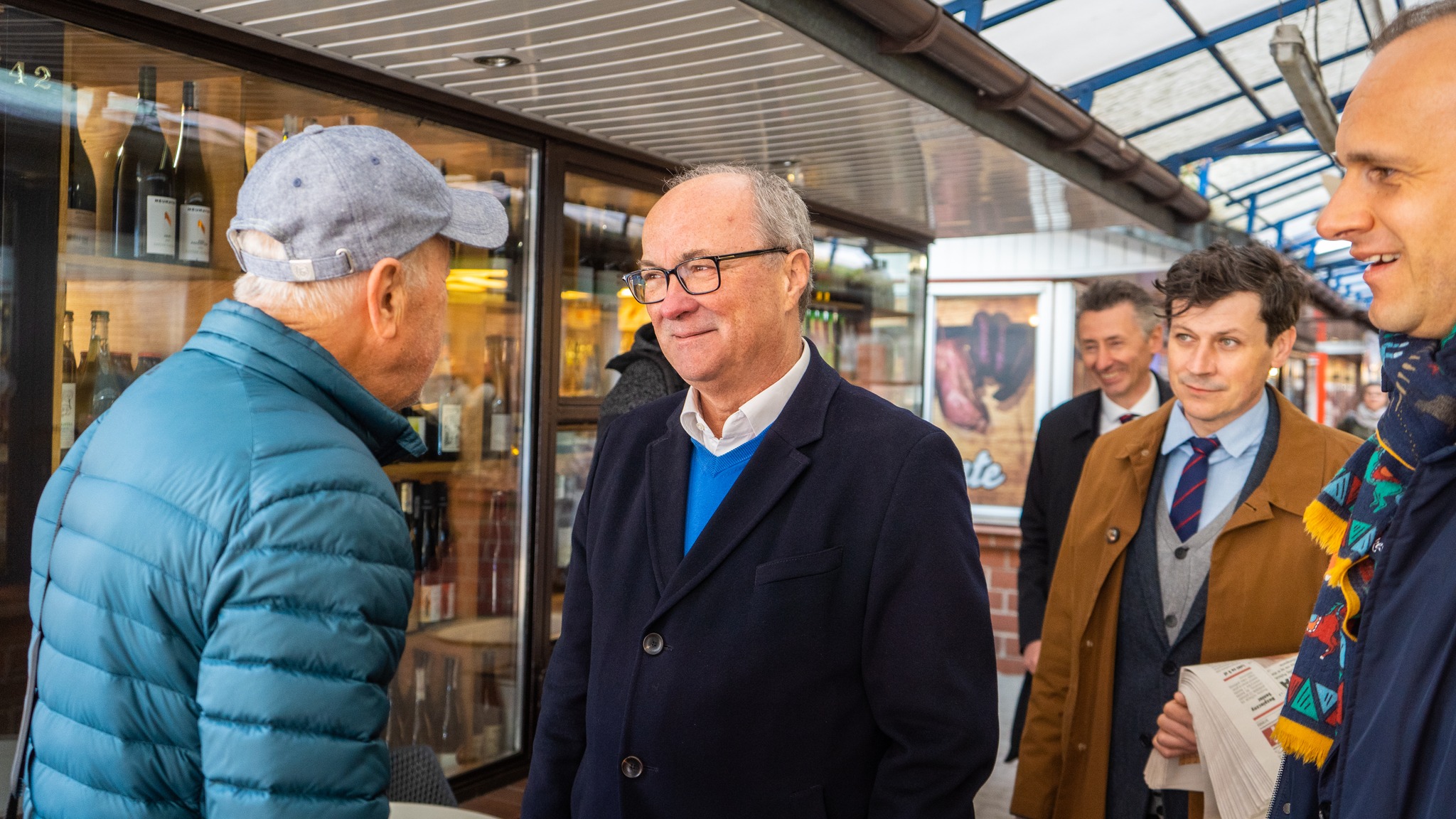 Grupa mężczyzn rozmawia w pasażu sklepowym z alkoholem; na pierwszym planie stoi uśmiechnięty mężczyzna w granatowym płaszczu i okularach, otoczony przez innych mężczyzn w kurtkach.