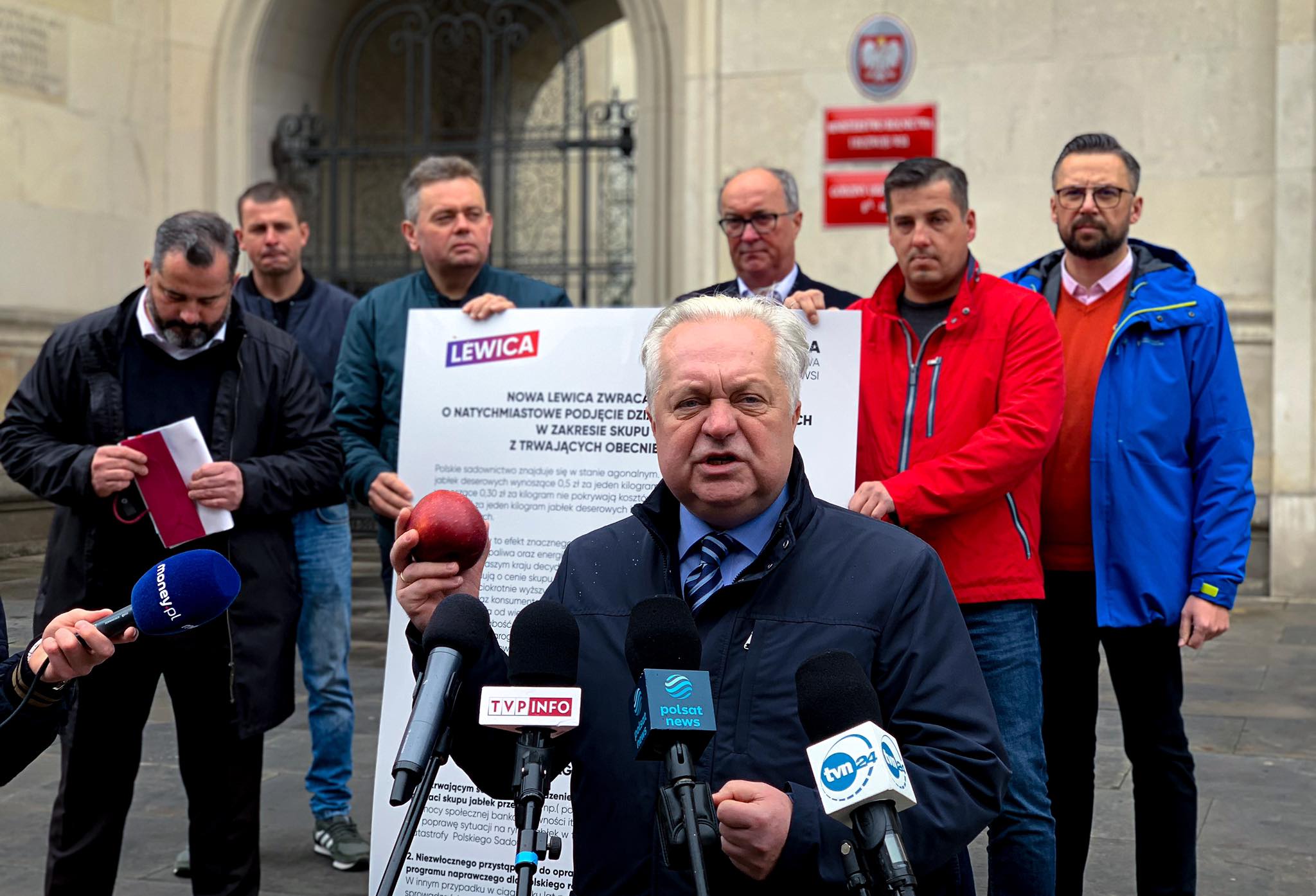 Polityk Lewicy przemawia na konferencji prasowej przed mikrofonami, stojąc obok grupy osób i plakatu z logiem Lewicy; temat wystąpienia to interwencyjny skup jabłek od polskich sadowników.