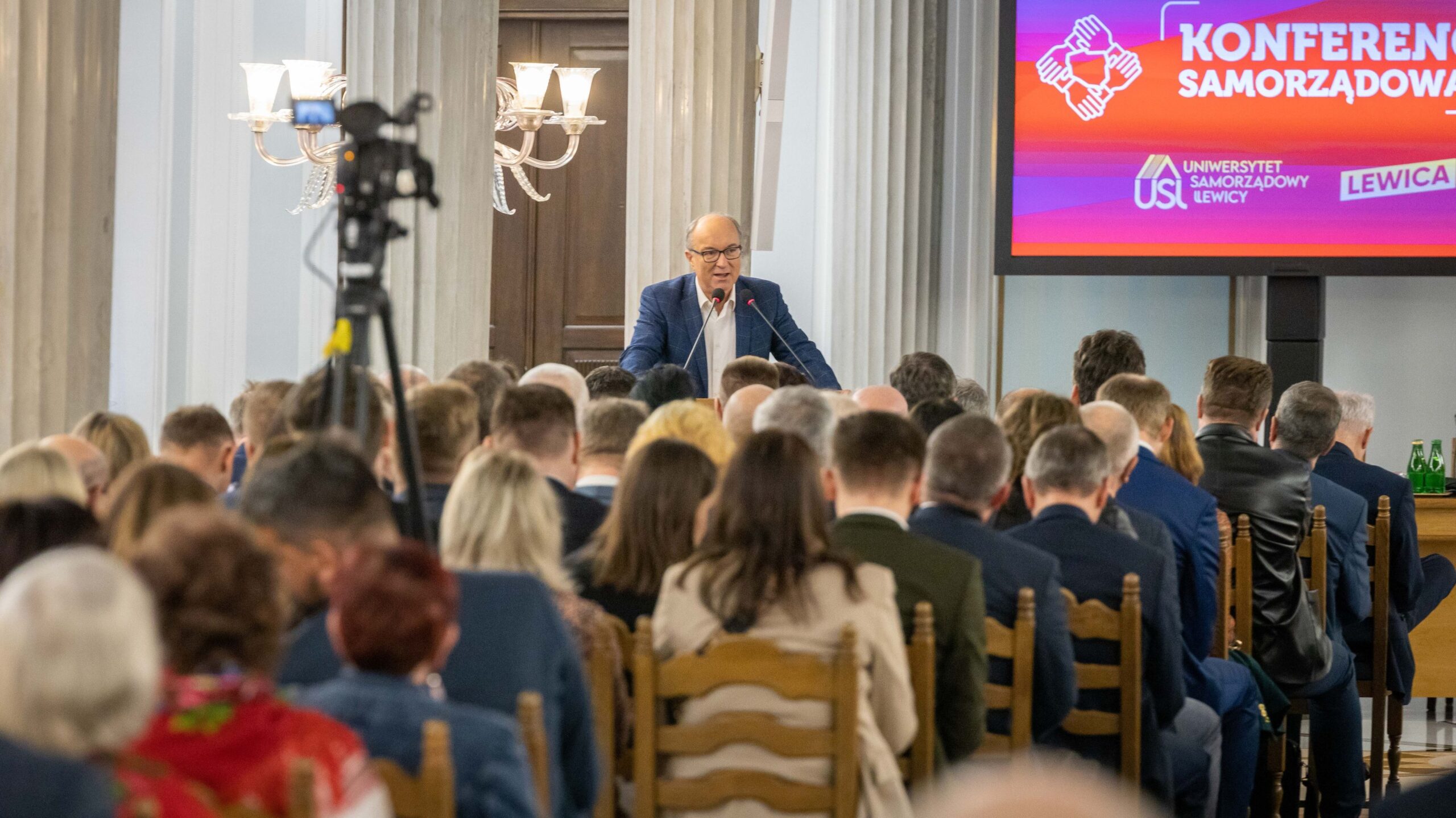 Na zdjęciu znajduje się mężczyzna przemawiający do zgromadzonego tłumu podczas konferencji samorządowej w eleganckim wnętrzu.