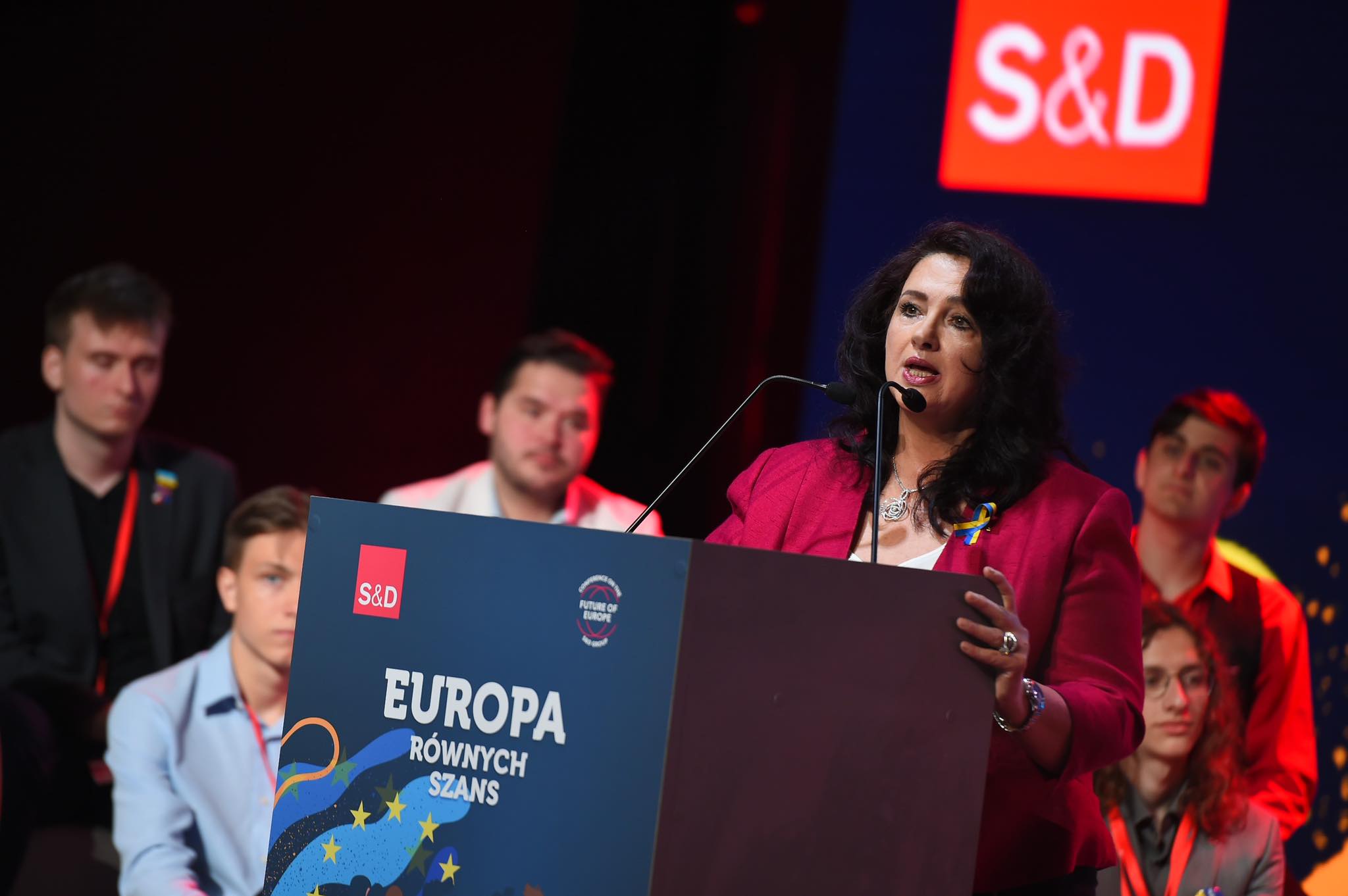 Na zdjęciu widoczna jest mówczyni stojąca przy mikrofonie na tle baneru z napisem