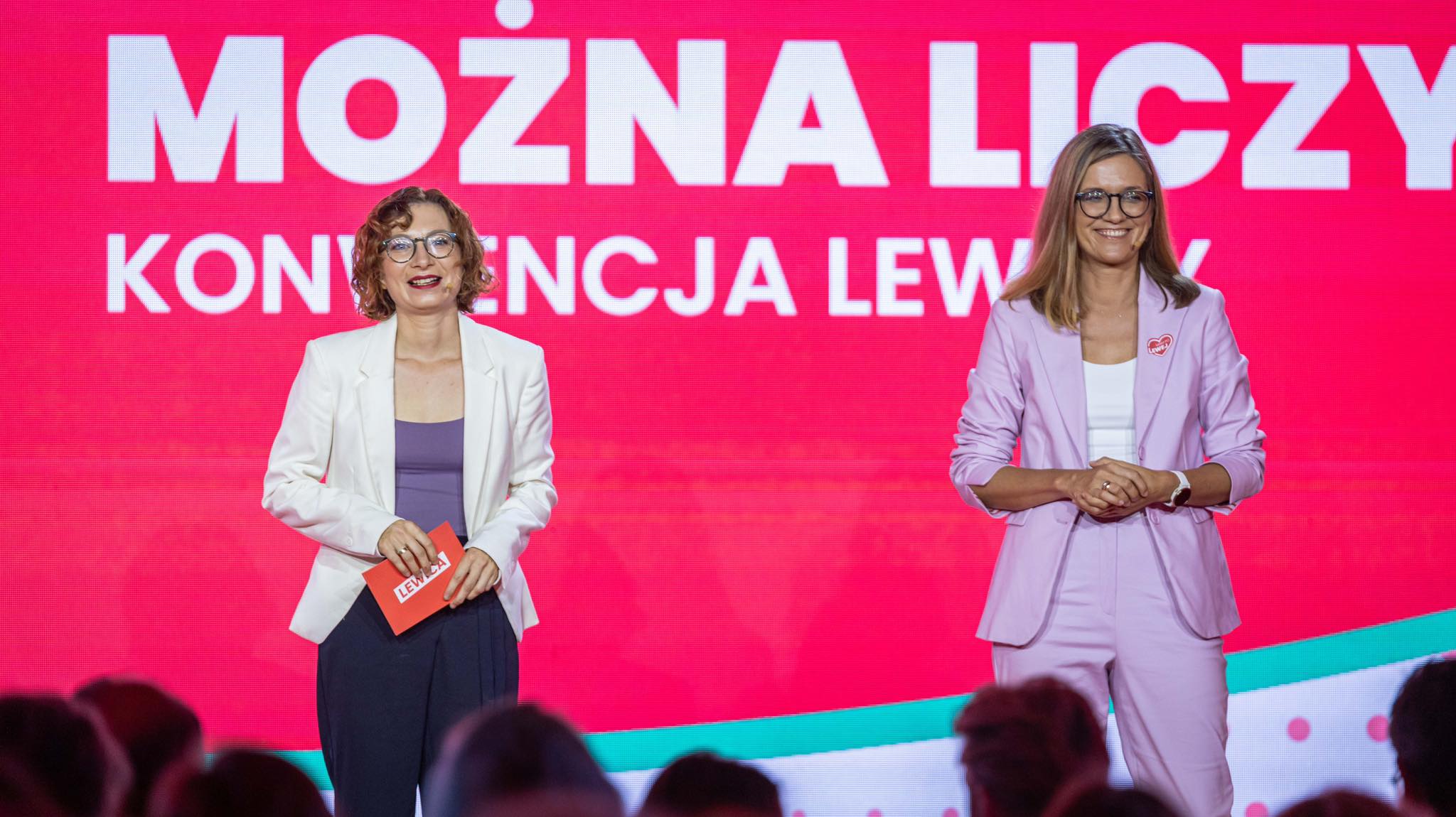 Dwie kobiety stoją na scenie podczas konferencji, na różowym tle z dużymi białymi napisami „MOŻNA LICZYĆ”, z czego jedna trzyma czerwoną kartkę z napisem Lewica.