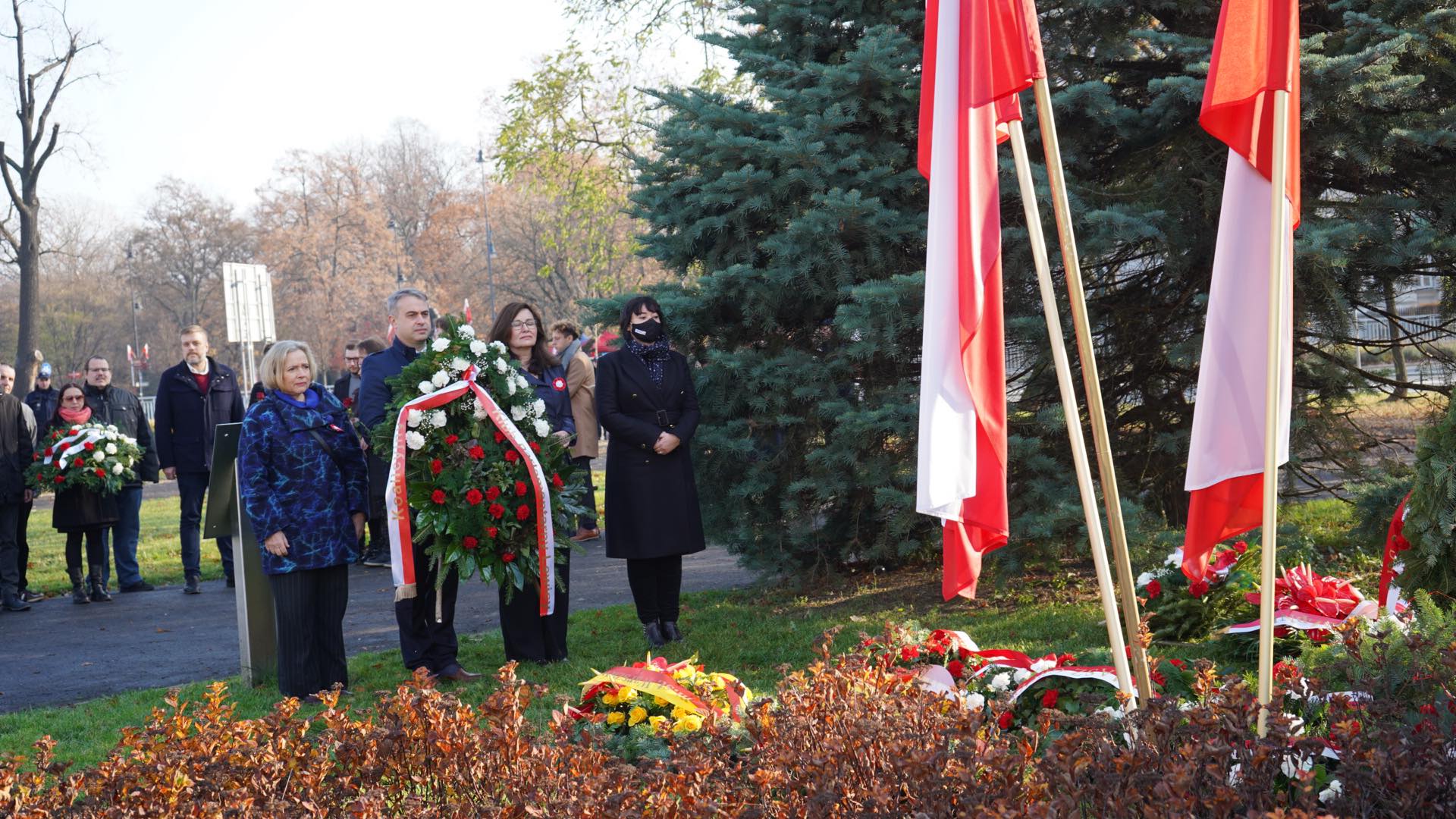 Grupa osób uczestniczy w uroczystości, składając wieńce w hołdzie, w otoczeniu drzew i flag narodowych.