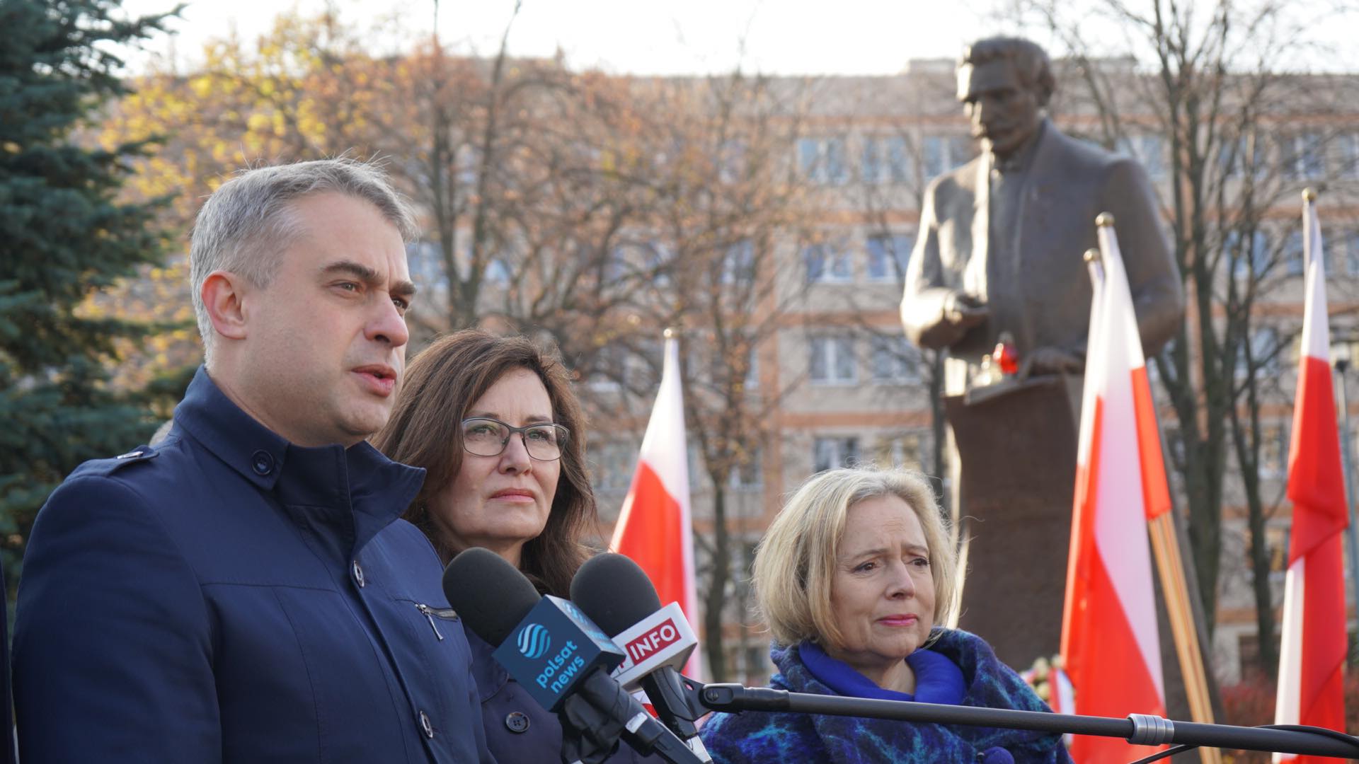 Trzy osoby udzielają konferencji prasowej podczas obchodów Narodowego Święta Niepodległości, z polskimi flagami w tle i posągiem widocznym za nimi.