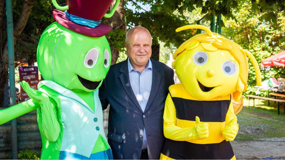 Mężczyzna w garniturze stoi między dwoma kolorowymi maskotkami w parku podczas plenerowego wydarzenia.