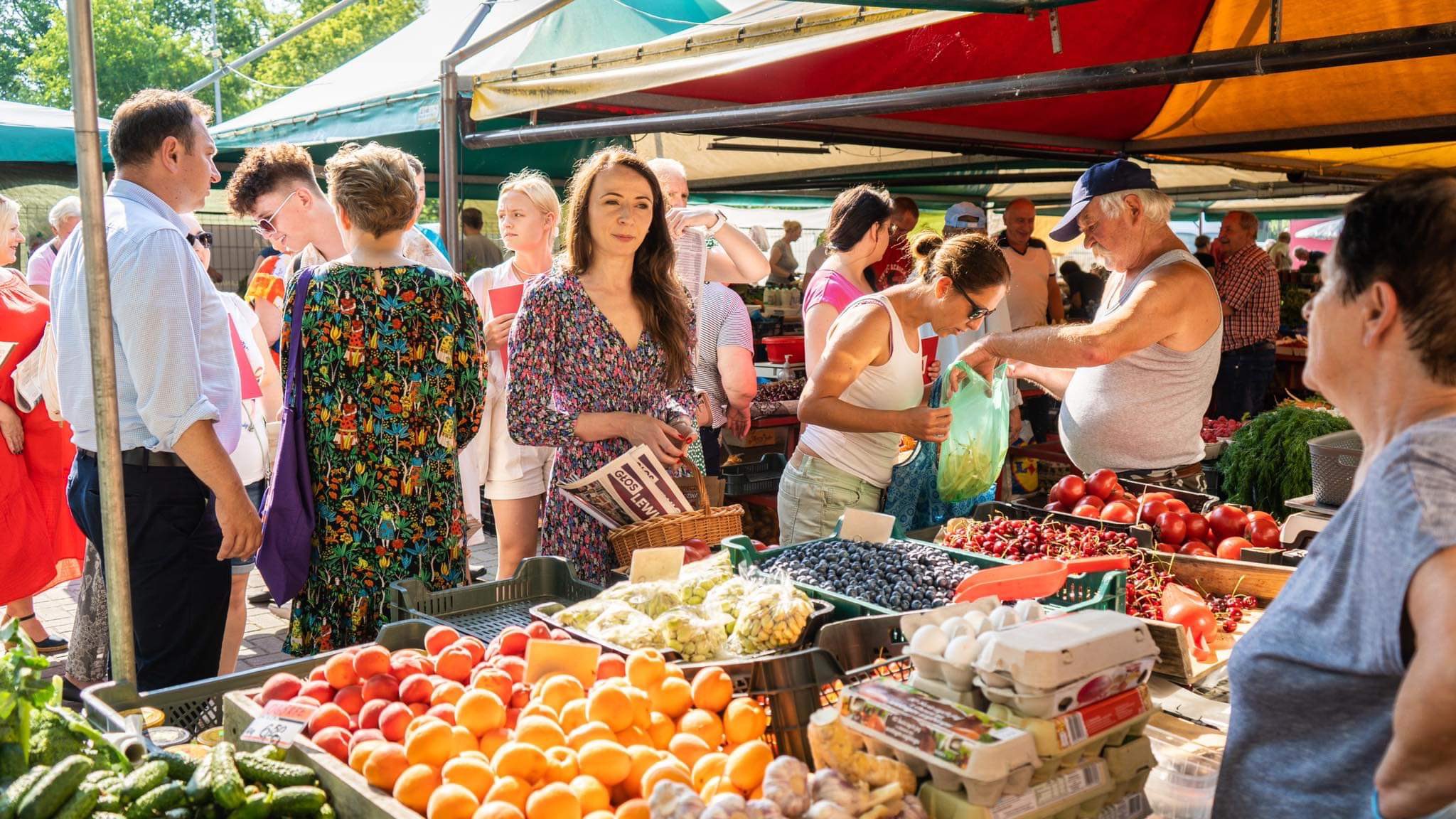 Grupa osób na targu spożywczym kupuje świeże owoce i warzywa pod kolorowymi parasolami, ilustrując codzienne zakupy w czasach rekordowej inflacji.