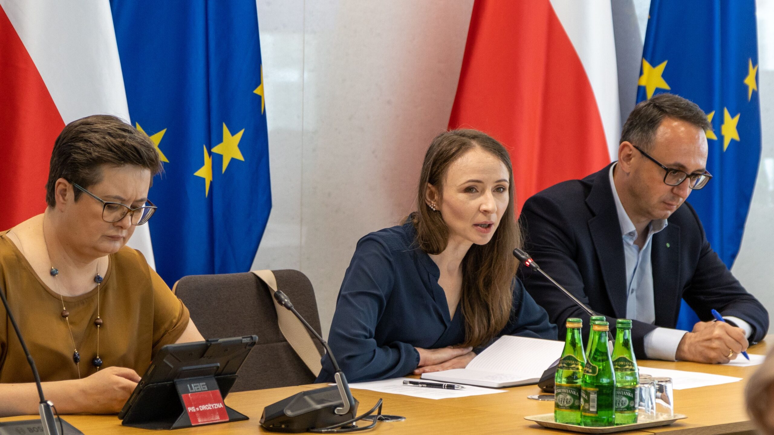 Na zdjęciu znajduje się spotkanie, podczas którego trzy osoby omawiają kwestie, siedząc przy stole, z flagami Polski i Unii Europejskiej w tle.