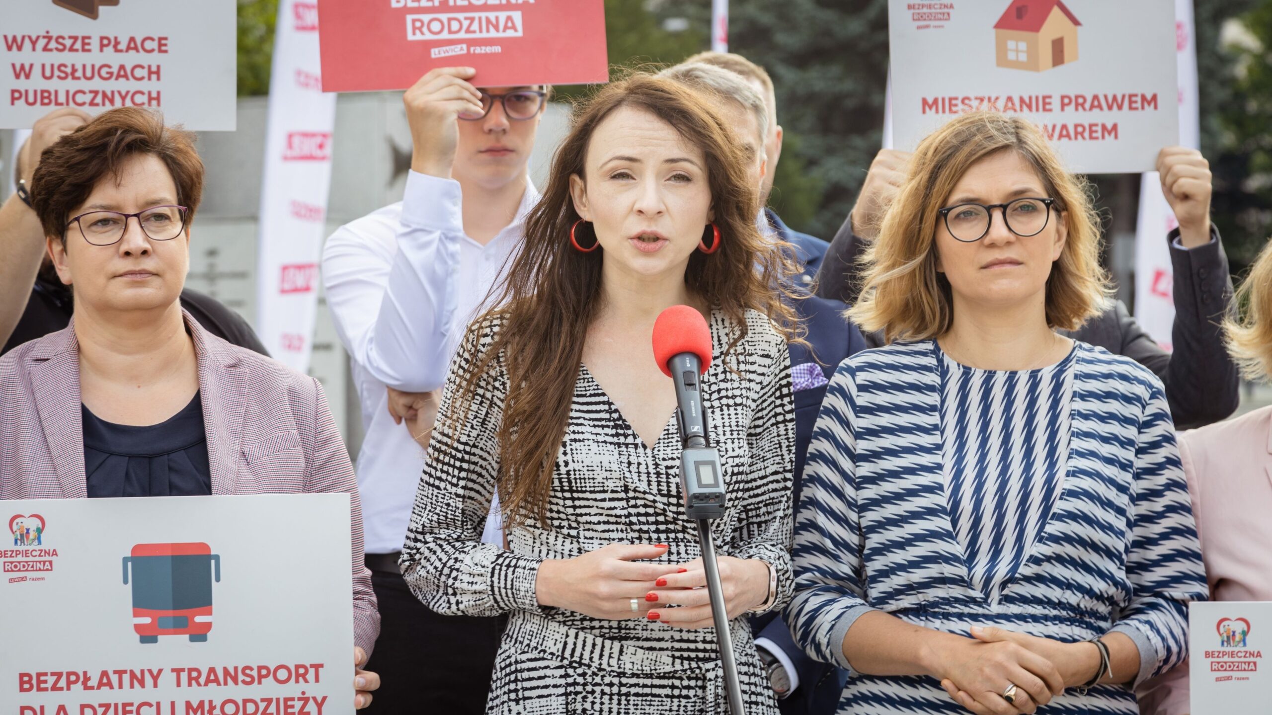 Na zdjęciu widać grupę ludzi podczas konferencji prasowej, gdzie jedna z kobiet przemawia do mikrofonu, a inni trzymają transparenty z hasłami dotyczącymi wsparcia rodzin i usług publicznych.