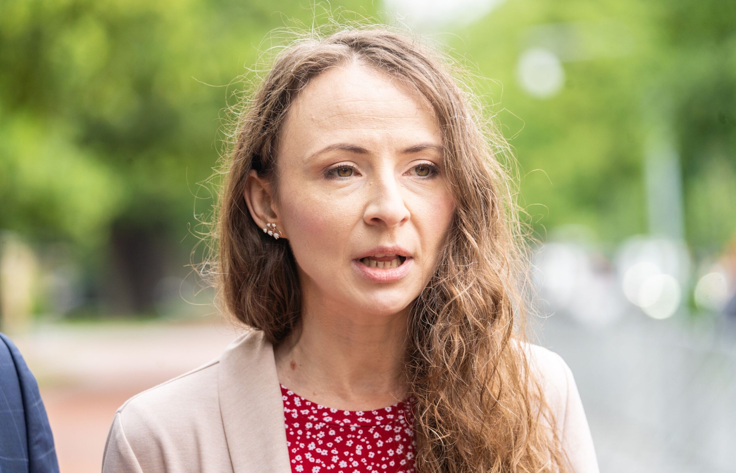 Na zdjęciu widać kobietę z długimi, kręconymi włosami, ubrana w czerwoną sukienkę w białe kwiaty oraz jasny żakiet, rozmawiającą na tle zielonego tła.