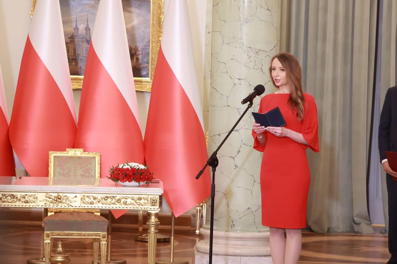 Kobieta w czerwonej sukience stoi przy mikrofonie, trzyma otwarty notatnik i przemawia podczas zaprzysiężenia rządu, w tle widoczne flagi Polski.