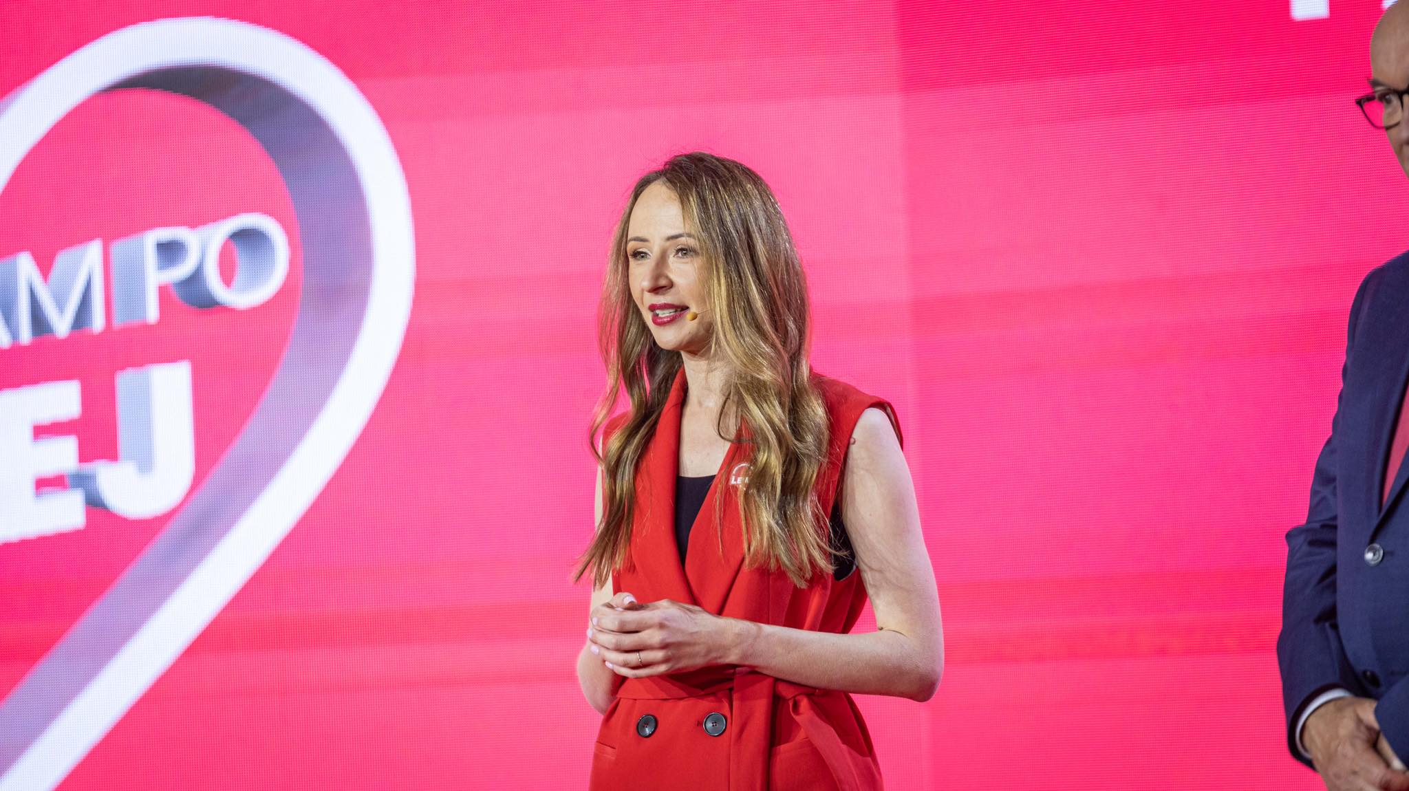 Kobieta z długimi blond włosami w czerwonym żakiecie przemawia na scenie przed różowym tłem z logo.