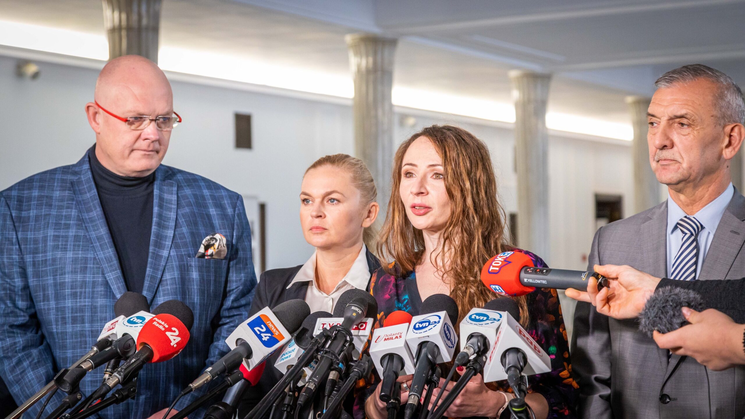 Na zdjęciu widoczna jest grupa czterech osób, które stoją z przodu mikrofonów podczas konferencji prasowej w jasnym wnętrzu.