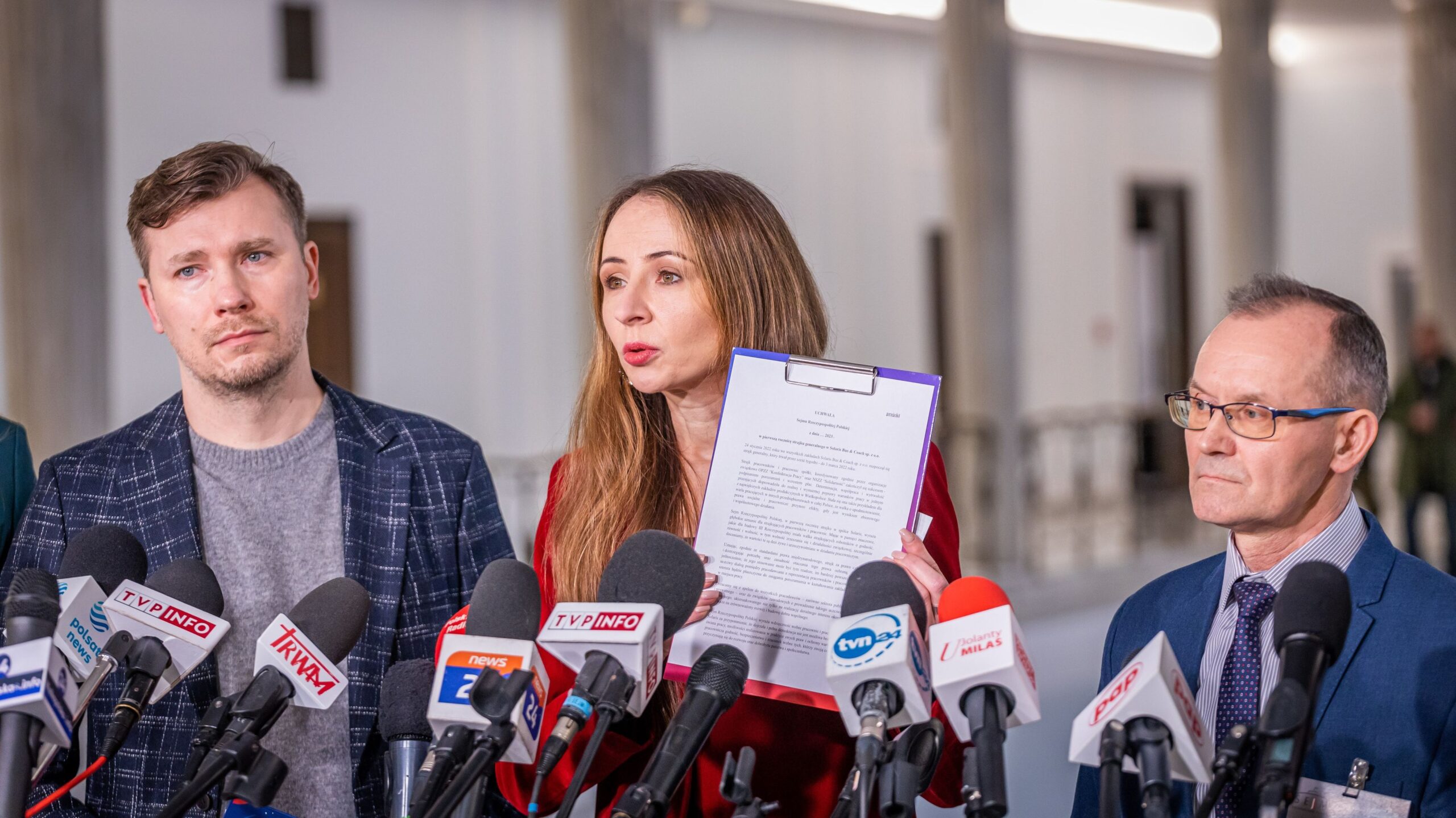 Na zdjęciu przedstawiono trzech ludzi podczas konferencji prasowej, z kobietą trzymającą dokument i mówiącą do mediów.