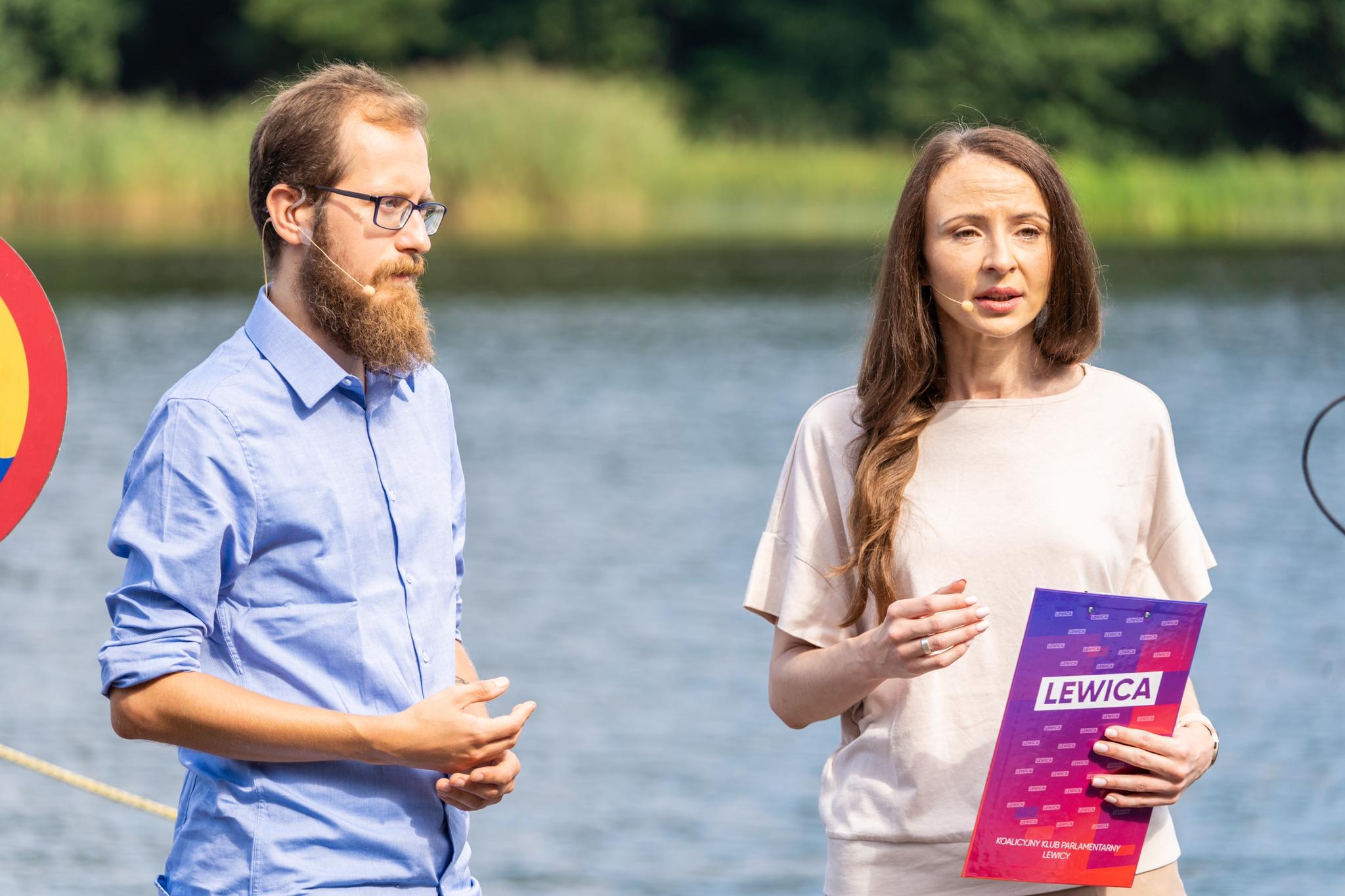 Para mówców przy jeziorze, mężczyzna z brodą i okularami oraz kobieta trzyma folder z logo LEWICA podczas konwencji programowej Lewicy.