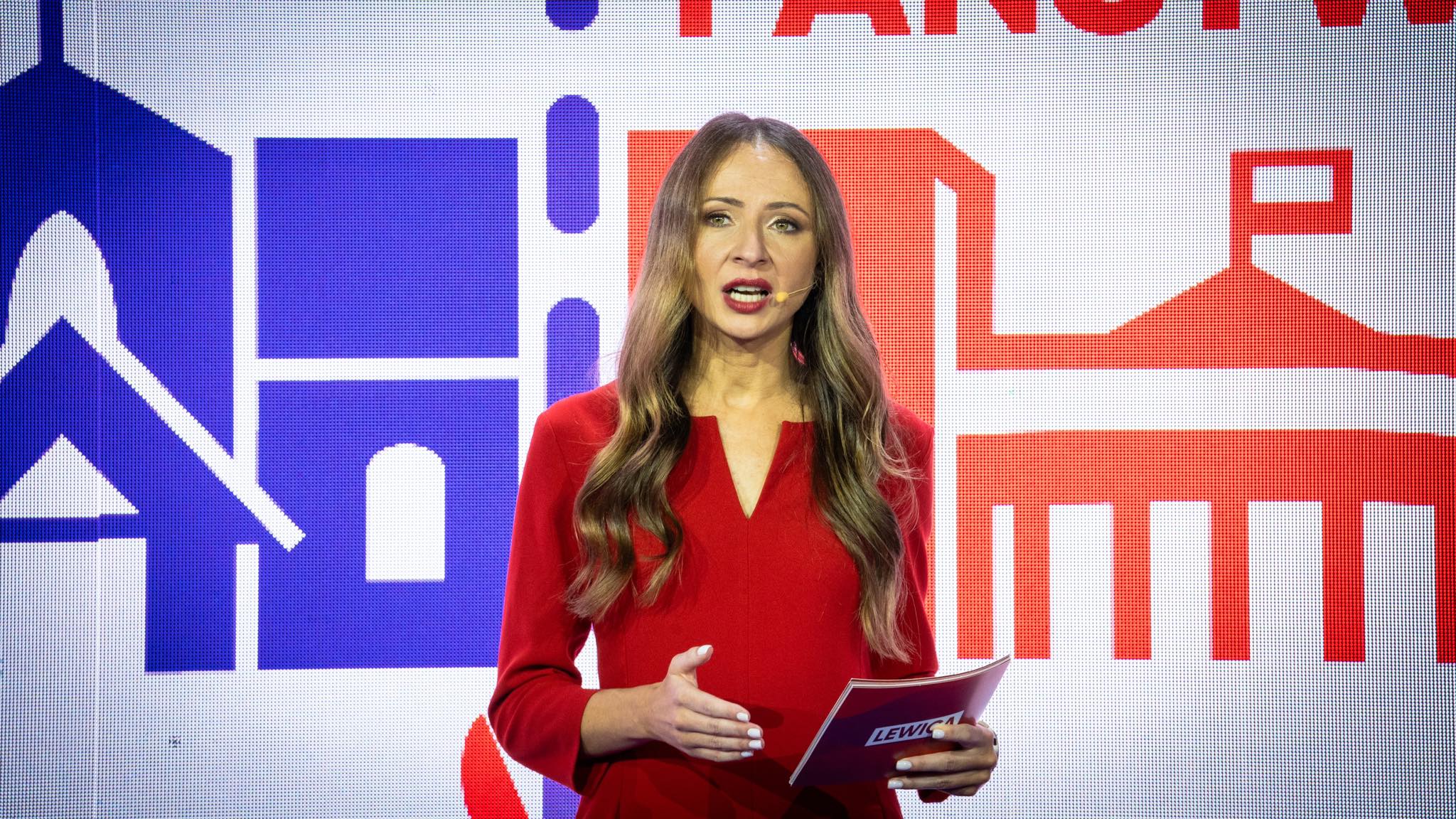 Prezenterka w czerwonej sukience z mikrofonem na twarzy stoi na scenie przed kolorowym, dużym tłem z grafikami i napisem „Świeckie państwo – Teraz!”.