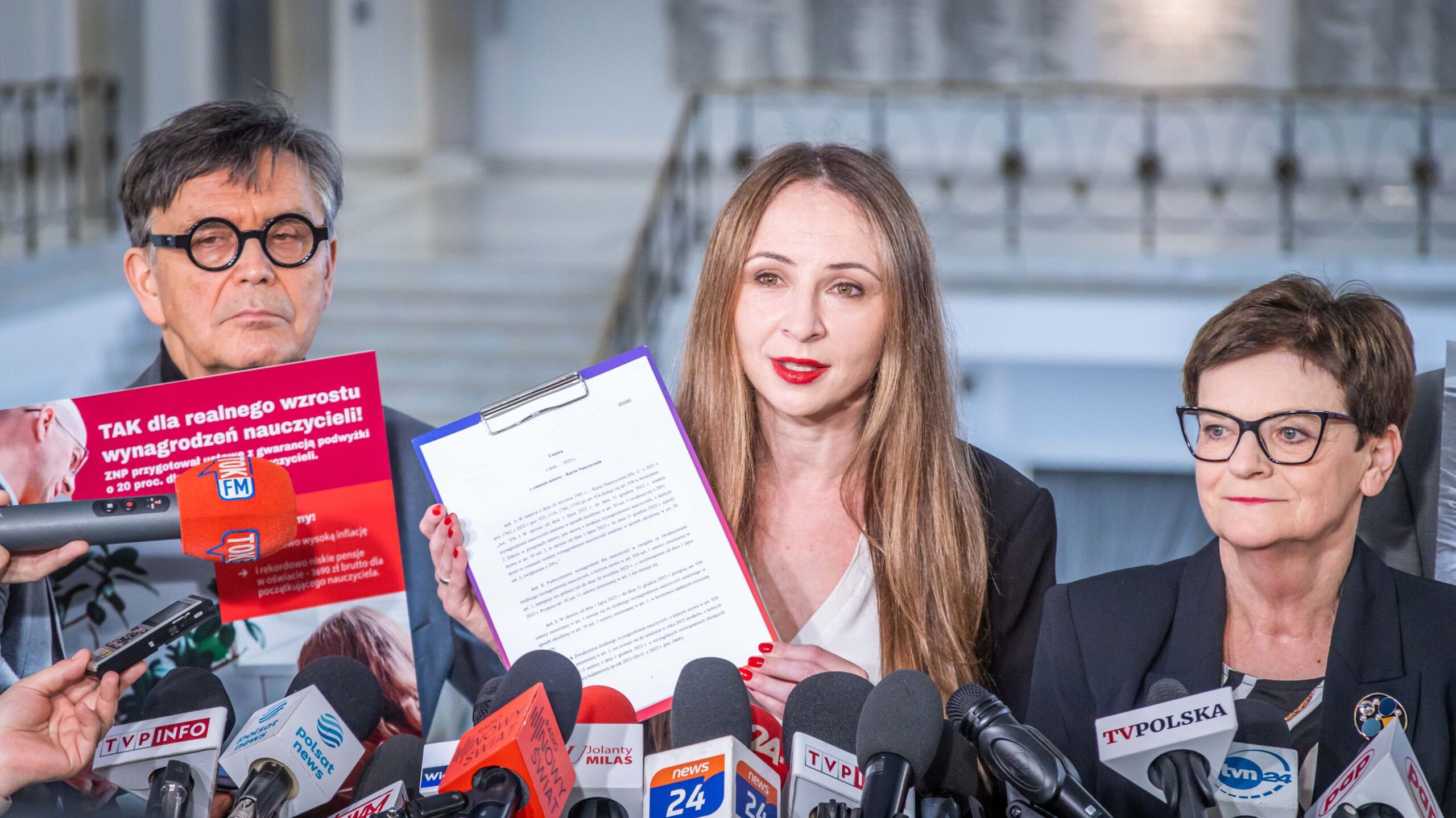 Na zdjęciu przedstawiono trzech mówców podczas konferencji prasowej, z których jedna osoba trzyma dokument, a w tle widać mikrofony i tabliczki z logo mediów.