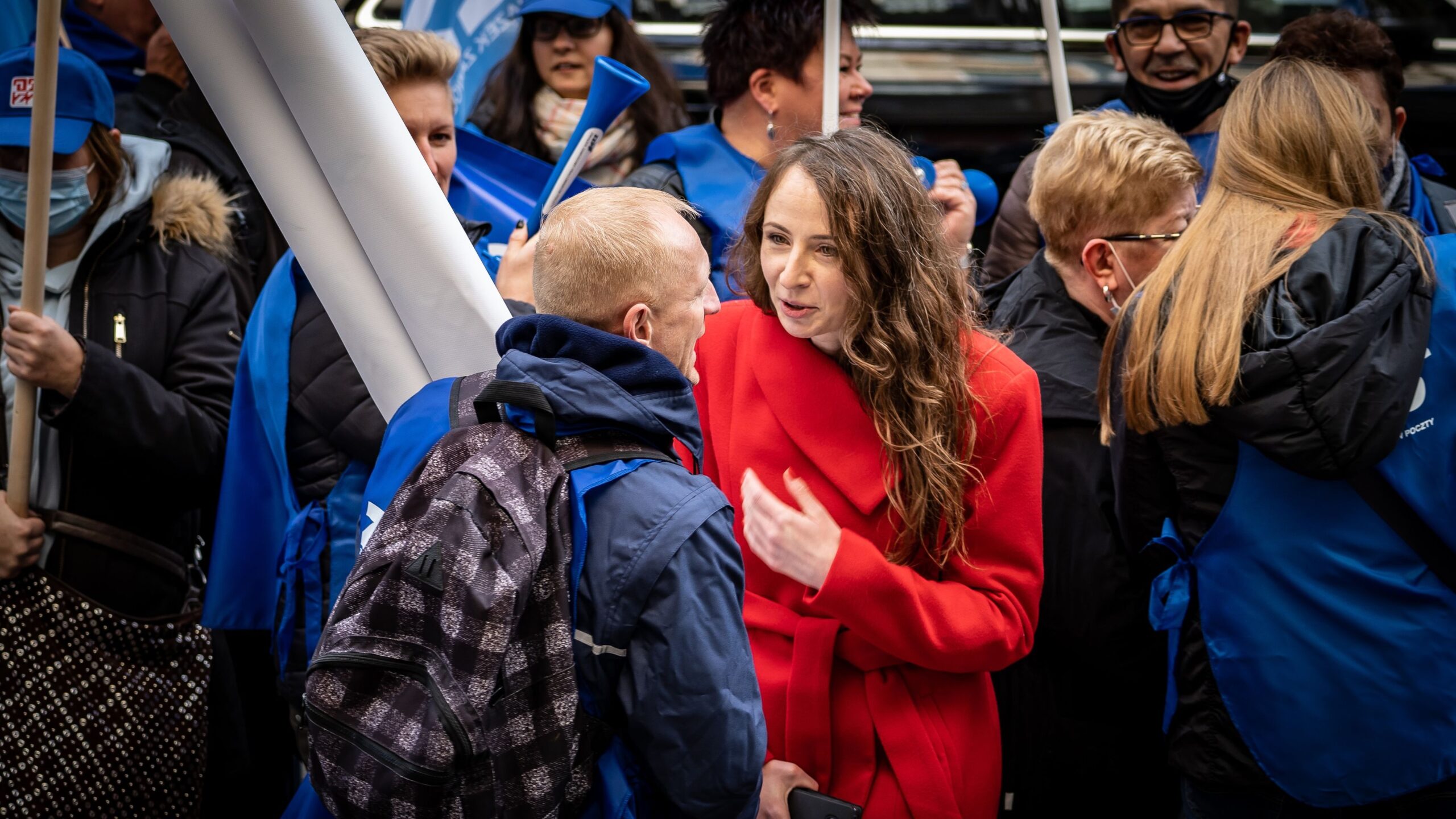 Kobieta w czerwonym płaszczu rozmawia z mężczyzną z plecakiem wśród tłumu uczestników demonstracji pracowników Poczty Polskiej.
