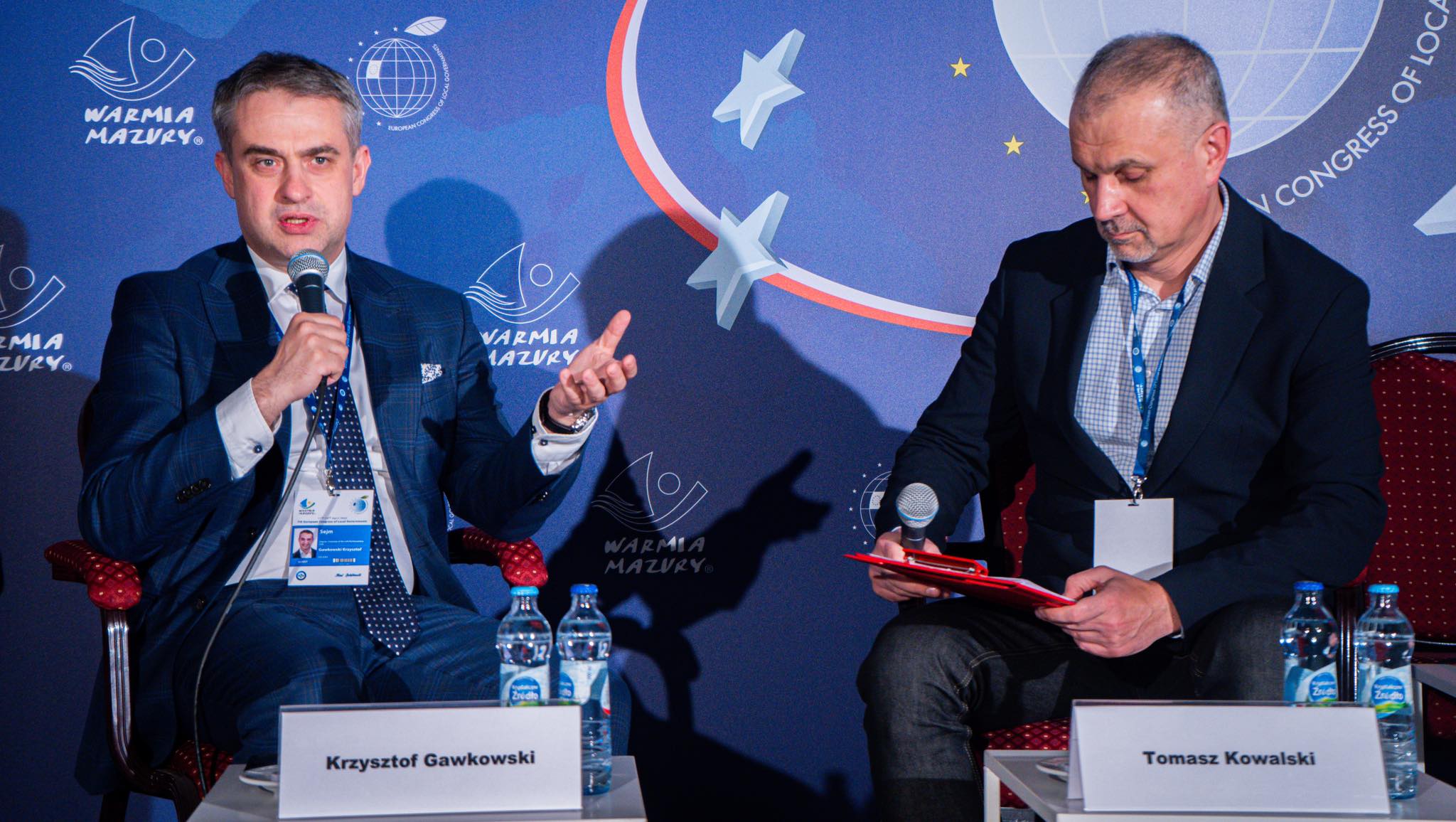 Dwóch mężczyzn siedzi na scenie podczas Europejskiego Kongresu Samorządów, jeden przemawia do mikrofonu, drugi trzyma notatki, a w tle widoczne są logotypy i grafika kongresu.