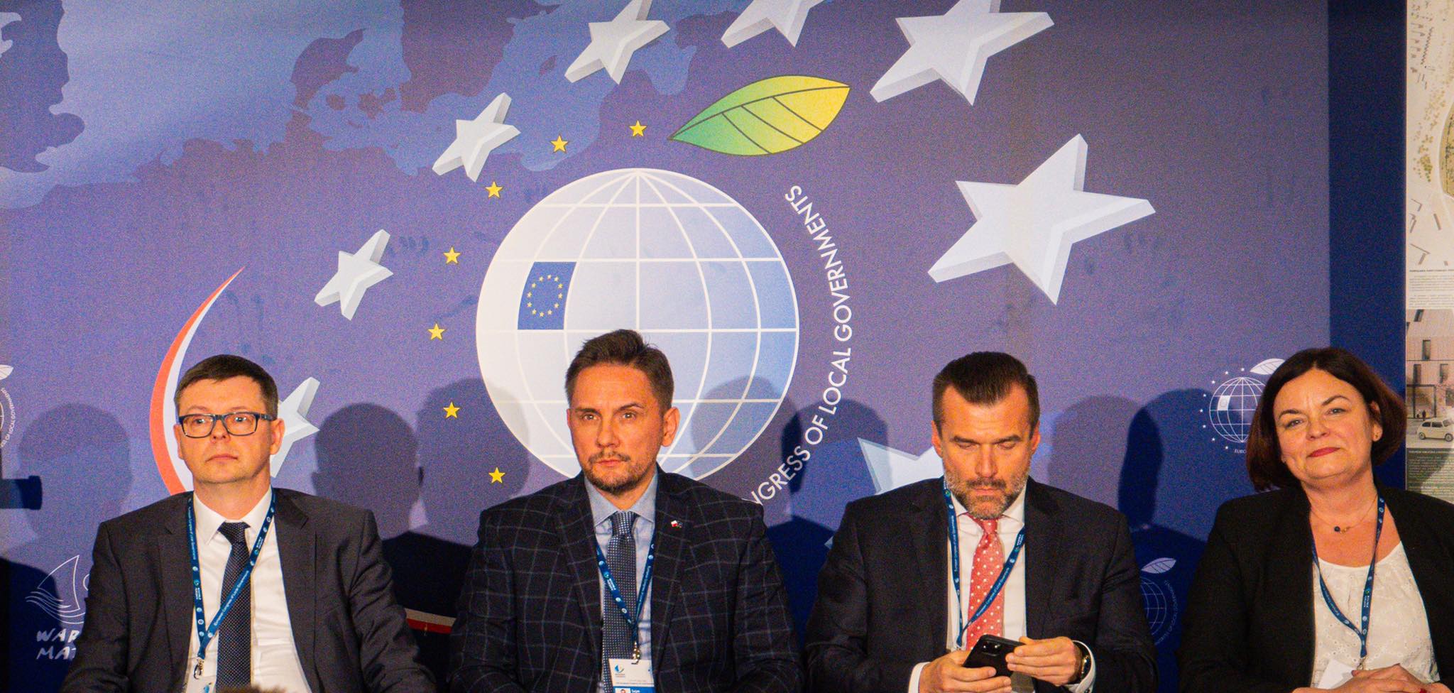 Czterech panelistów w garniturach siedzi na scenie przed niebieskim tłem z logo Europejskiego Kongresu Samorządów.