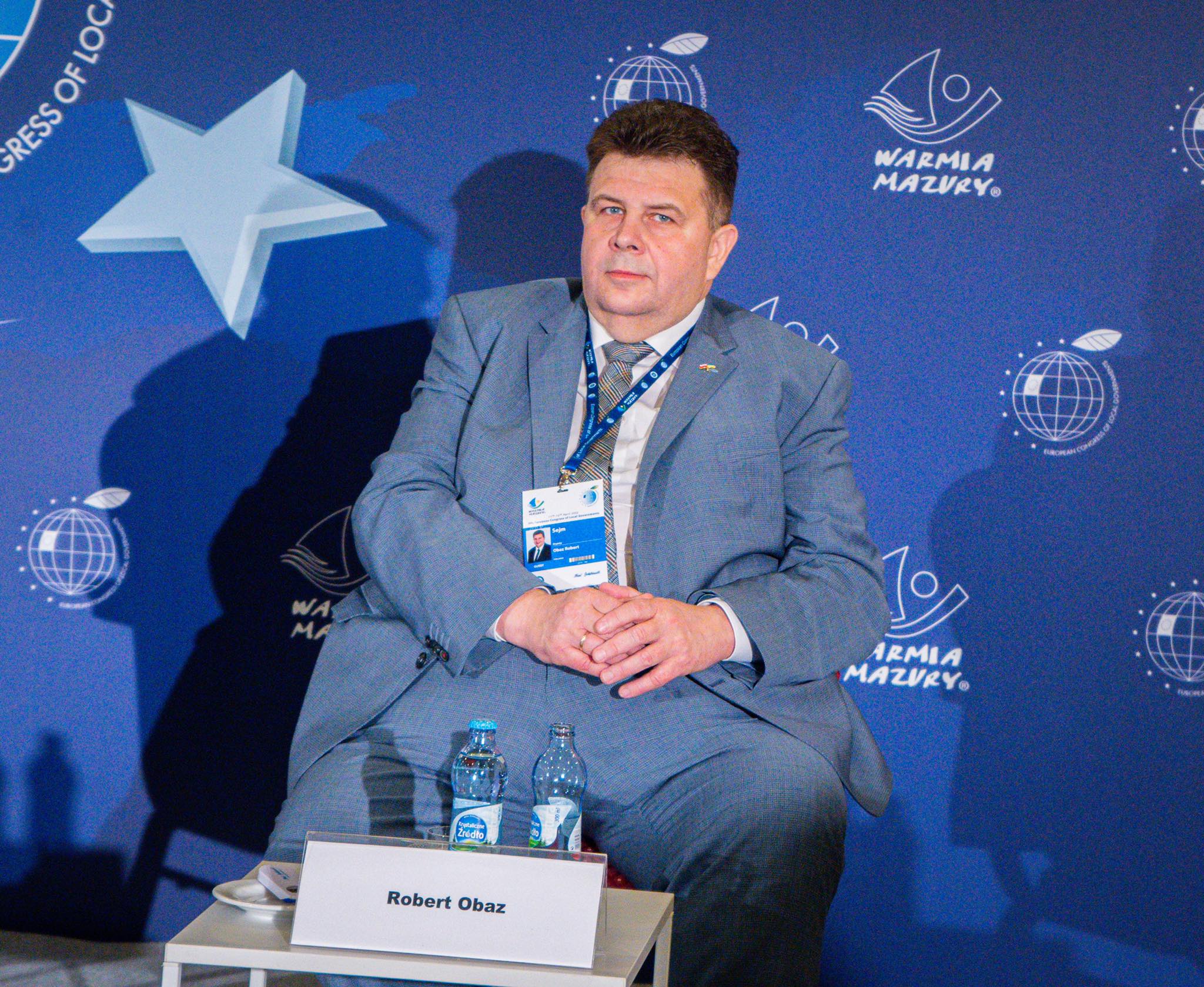 Mężczyzna w garniturze siedzi na scenie podczas Europejskiego Kongresu Samorządów, na tle niebieskiego tła z logotypami, z identyfikatorem na szyi i dwoma butelkami wody na stoliku przed nim.