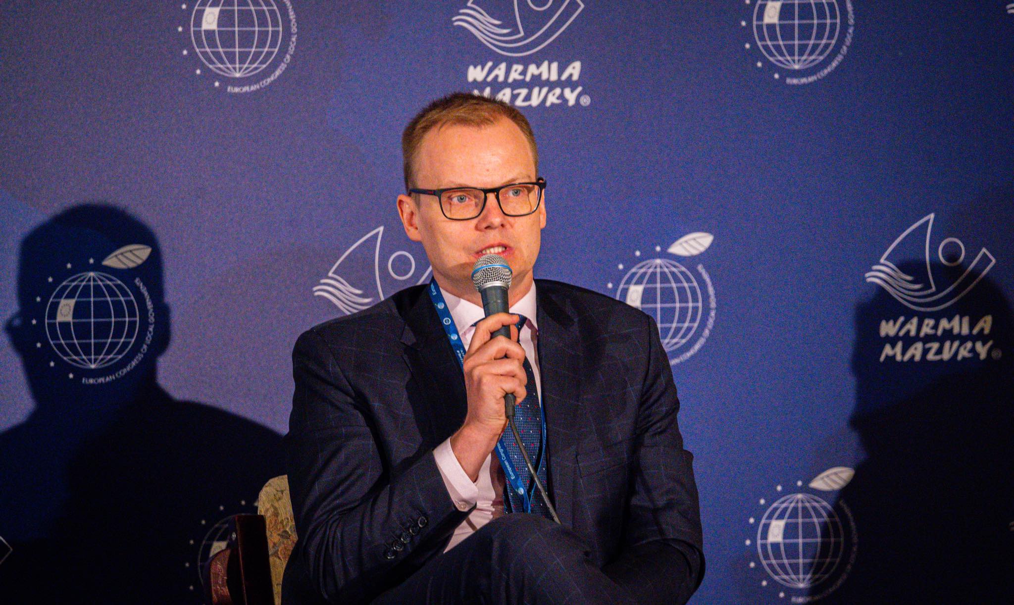 Mężczyzna w garniturze z okularami przemawia do mikrofonu podczas Europejskiego Kongresu Samorządów, na niebieskim tle z logo Warmia Mazury.