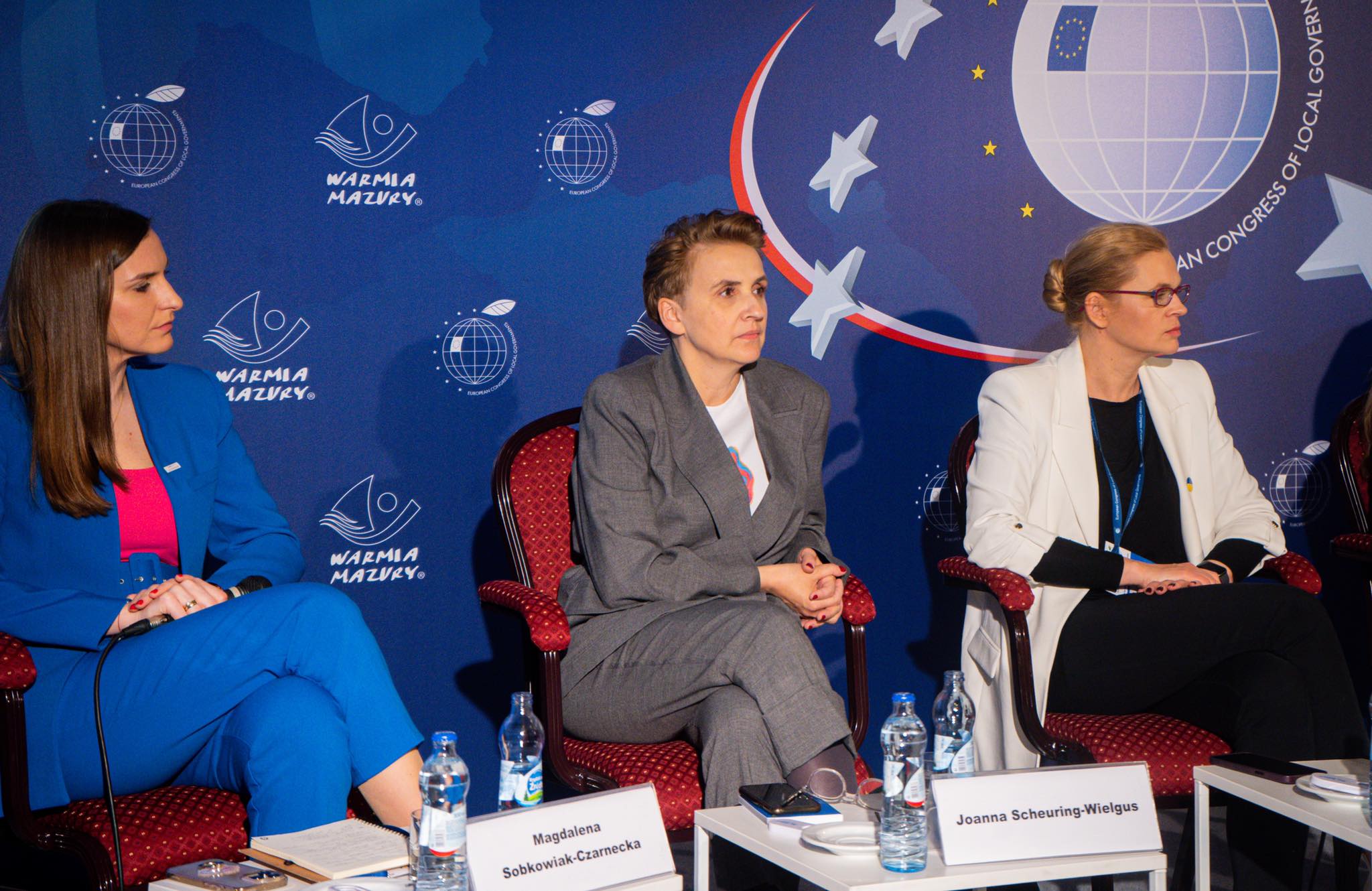 Trzy kobiety siedzą na panelu podczas Europejskiego Kongresu Samorządów, przed niebieską ścianką z logotypami i globem, z butelkami wody i notesami na stołach.