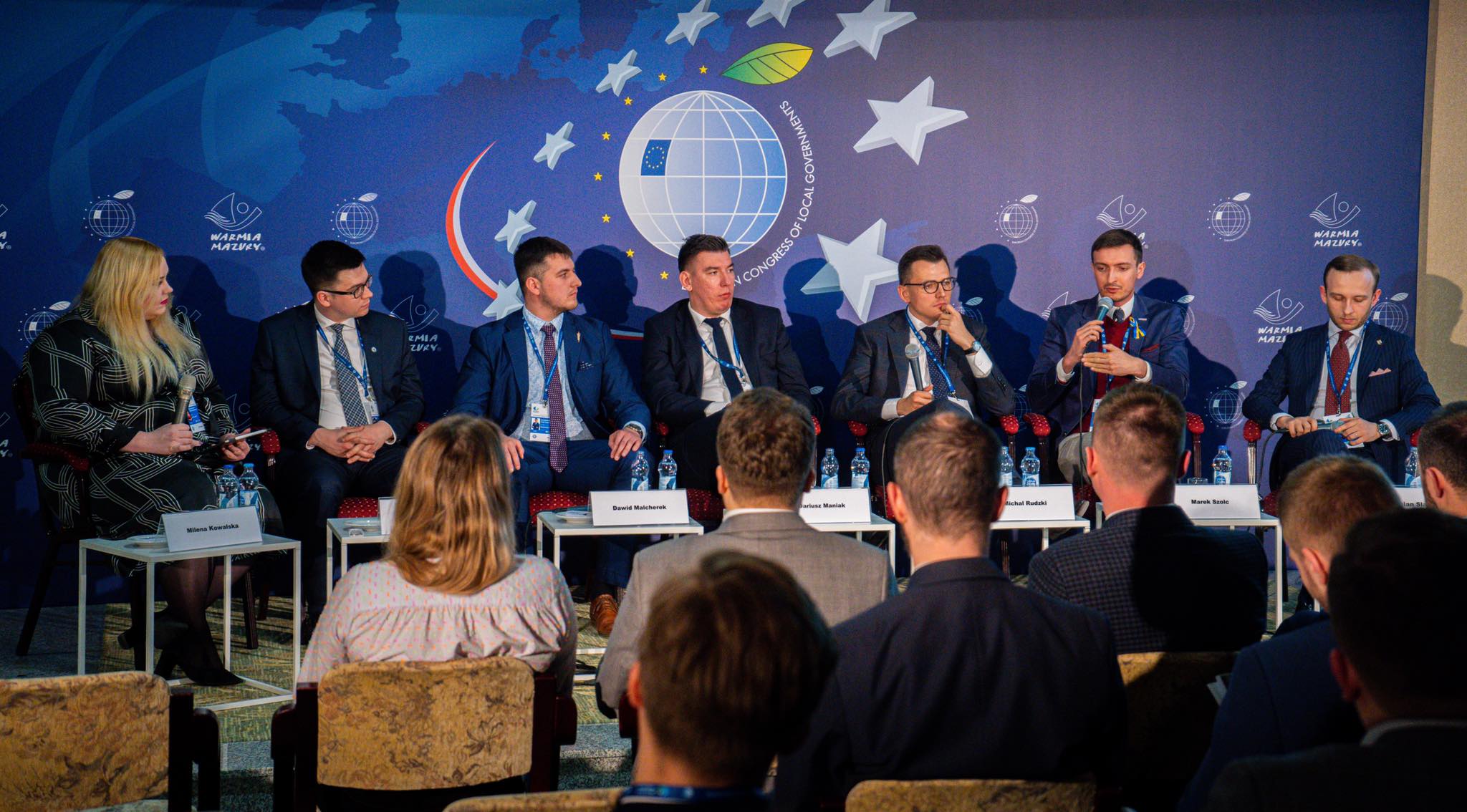 Uczestnicy konferencji siedzą za stołami, dyskutując na temat lokalnych rządów przed tłem z motywem mapy Europy i symbolami Unii Europejskiej.