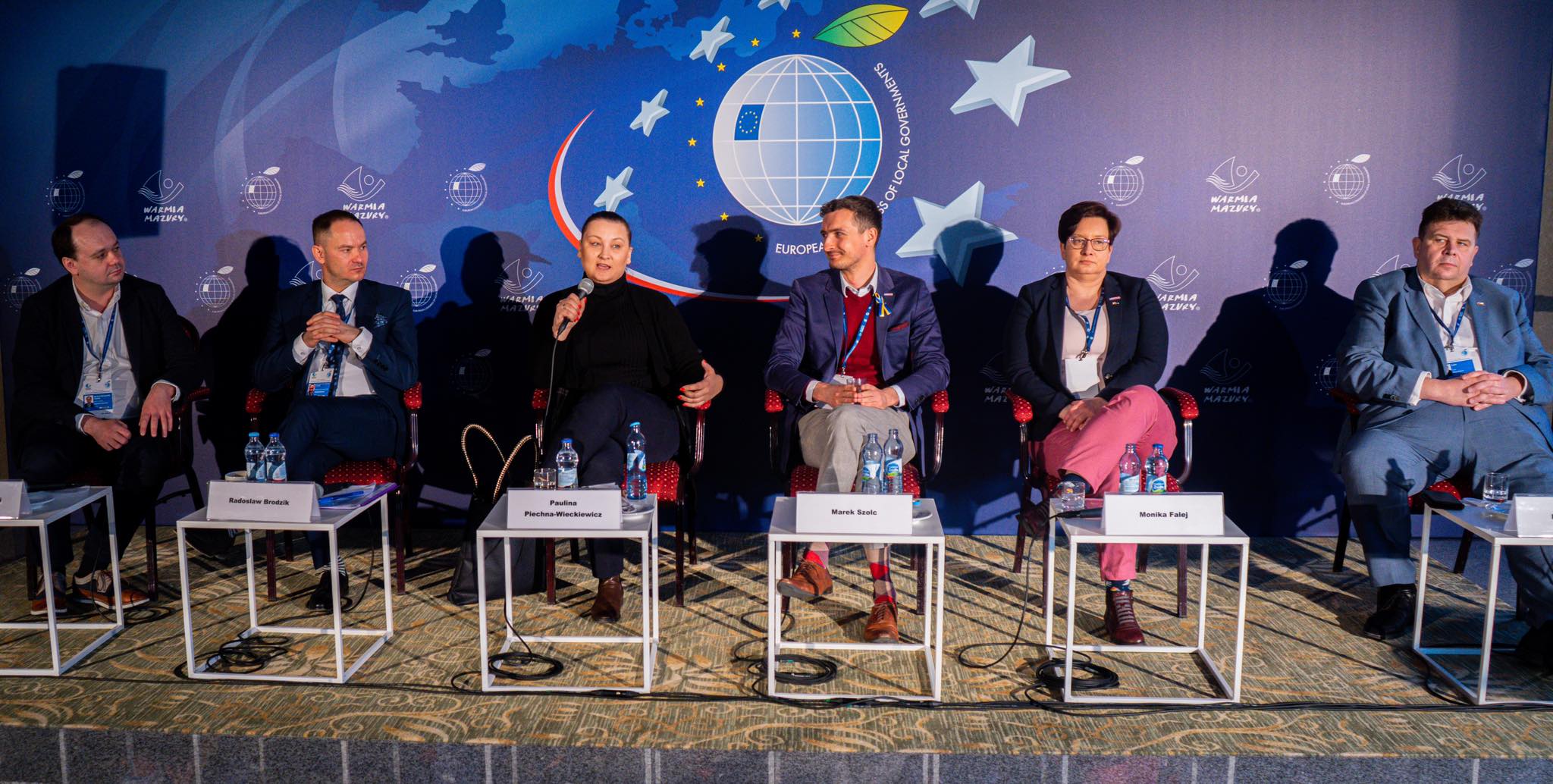 Szóstka panelistów siedzi na scenie podczas debaty na Europejskim Kongresie Samorządów, przed nimi stoliki z wodą i identyfikatorami, a w tle widoczne jest niebieskie tło z motywem globu, gwiazd i logotypem kongresu.