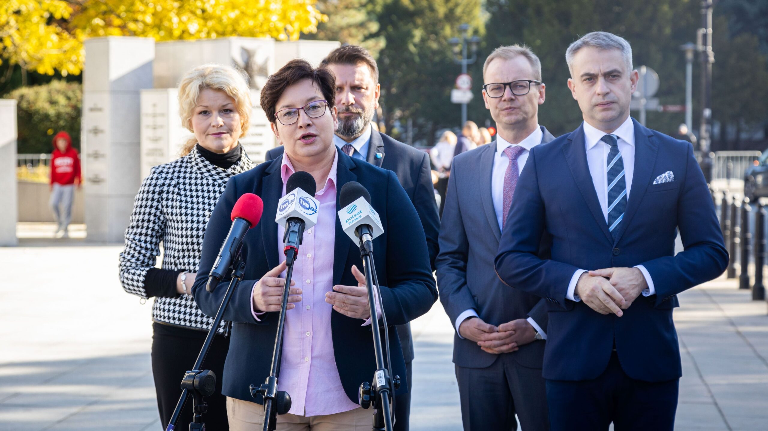 Na zdjęciu widoczna jest grupa polityków podczas konferencji prasowej na tle jesiennego krajobrazu.