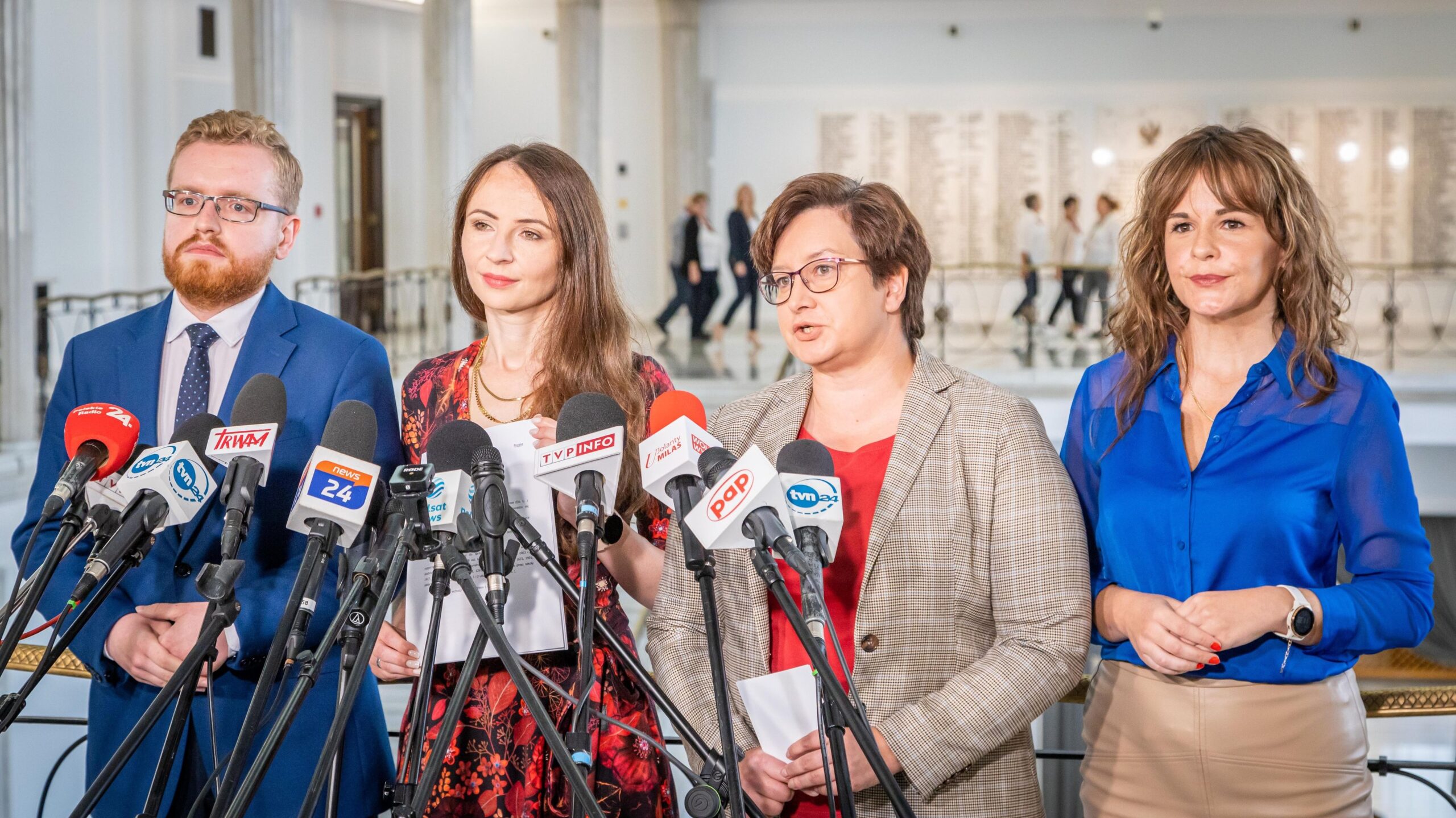 Na zdjęciu cztery osoby na konferencji prasowej stoją przed mikrofonami w budynku publicznym.