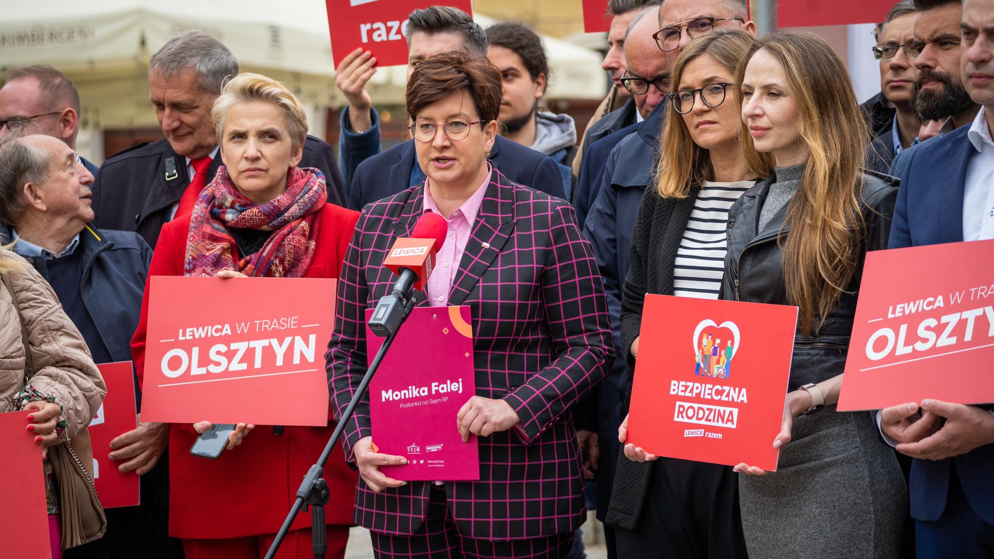 Grupa zwolenników Lewicy na wiecu na Warmii i Mazurach w Olsztynie, z kobietą w różowej kratkowanej marynarce przy mikrofonie i czerwonymi plakatami.