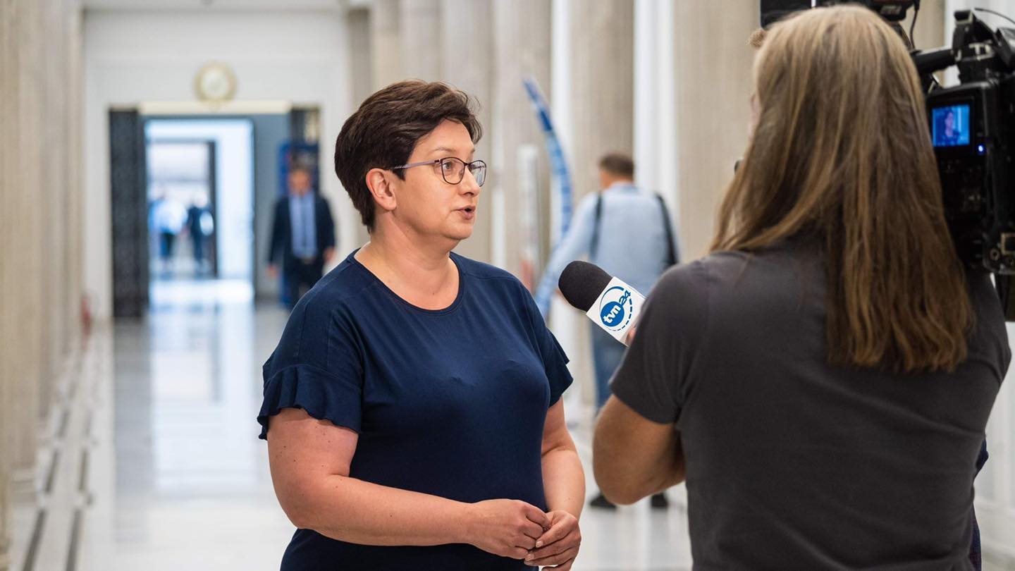 Na zdjęciu widać kobietę w granatowej koszulce, udzielającą wywiadu przed kamerą w przestronnym korytarzu.