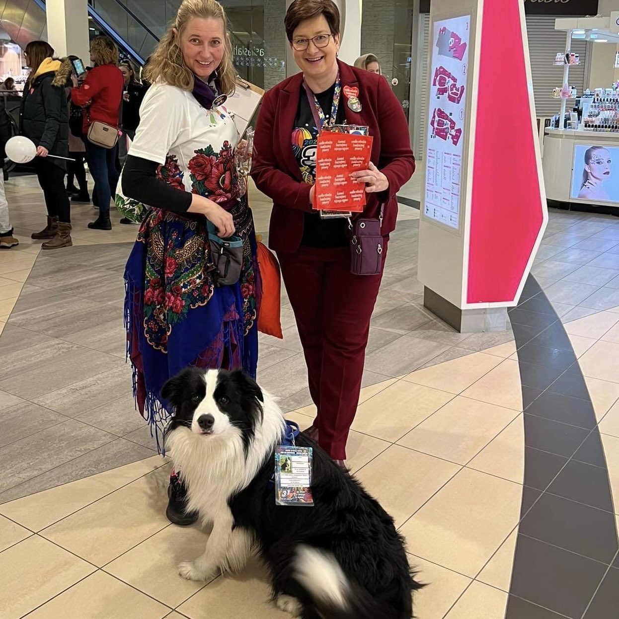 Dwie kobiety w galerii handlowej podczas WOŚP 2023 (Gramy do końca świata i jeden dzień dłużej), jedna w kwiecistej białej bluzce, druga w bordo, trzymają czerwone ulotki, a przed nimi siedzi czarno-biały border collie z identyfikatorem.