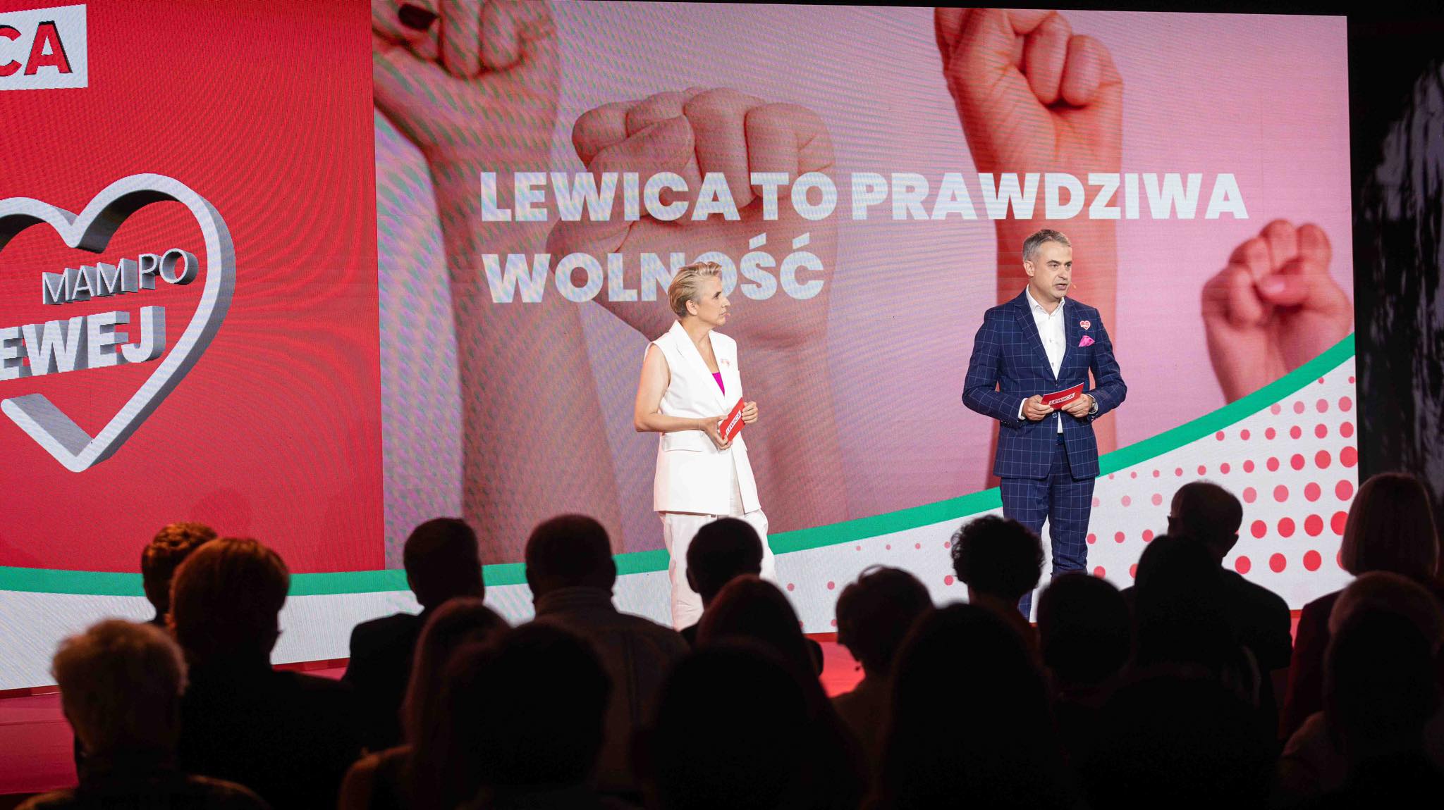 Dwóch prowadzących na scenie podczas wydarzenia politycznego, stoją przed dużym ekranem z napisem „LEWICA TO PRAWDZIWA WOLNOŚĆ” i kolorowym tłem, przed zgromadzoną publicznością.