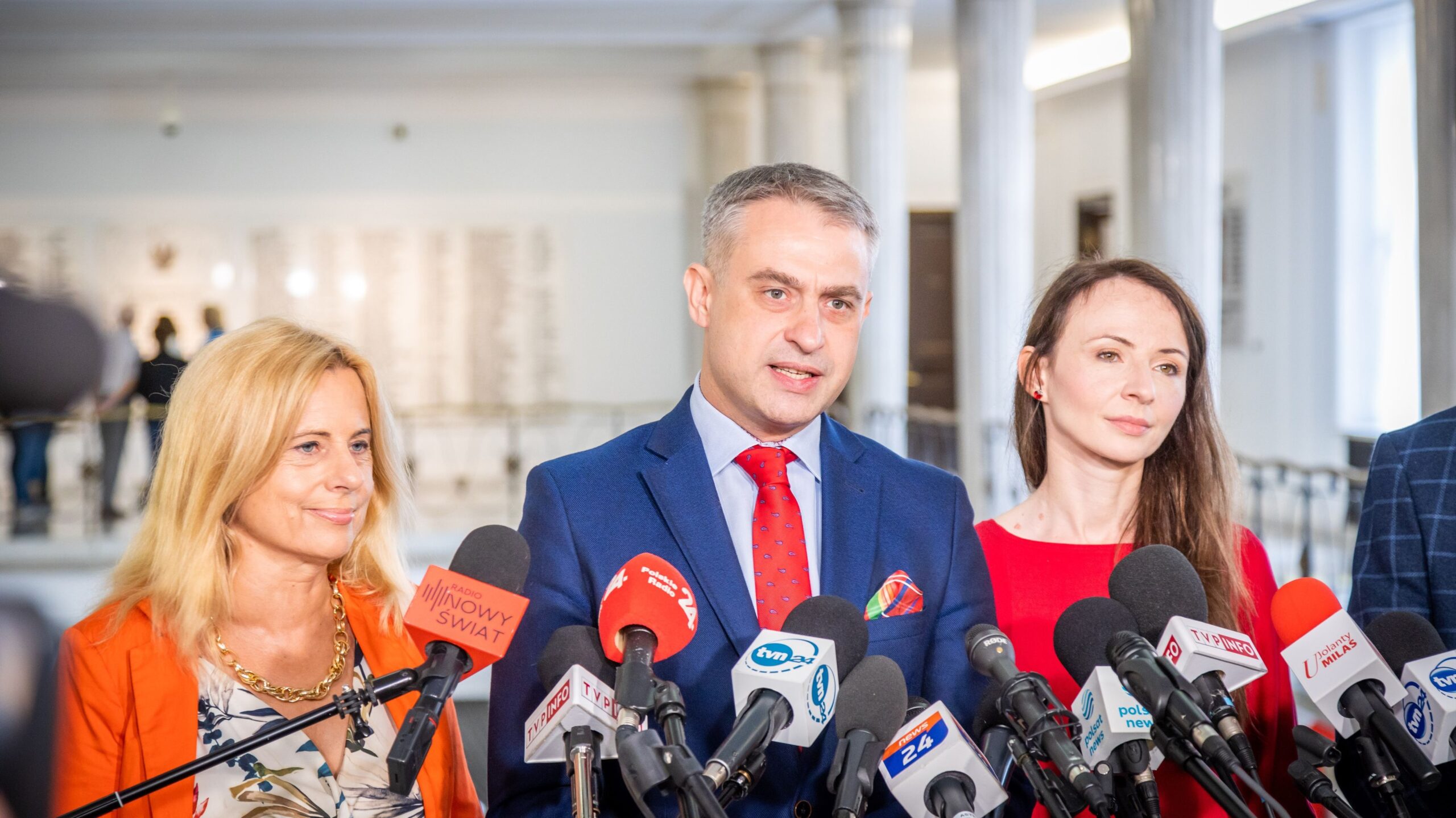 Na zdjęciu widać grupę trzech osób, w tym mężczyznę w garniturze oraz dwie kobiety, stojących przed mikrofonami i udzielających informacji w miejscu publicznym.