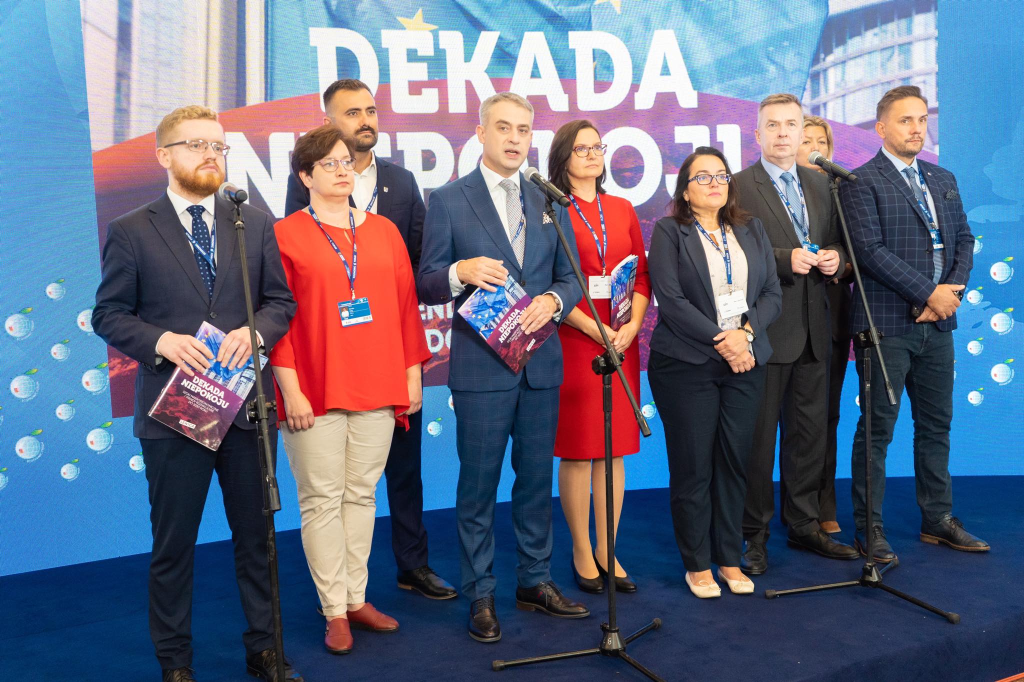 Na zdjęciu grupa ludzi stoi na scenie, trzymając dokumenty i mówiąc do mikrofonów, podczas wydarzenia o nazwie