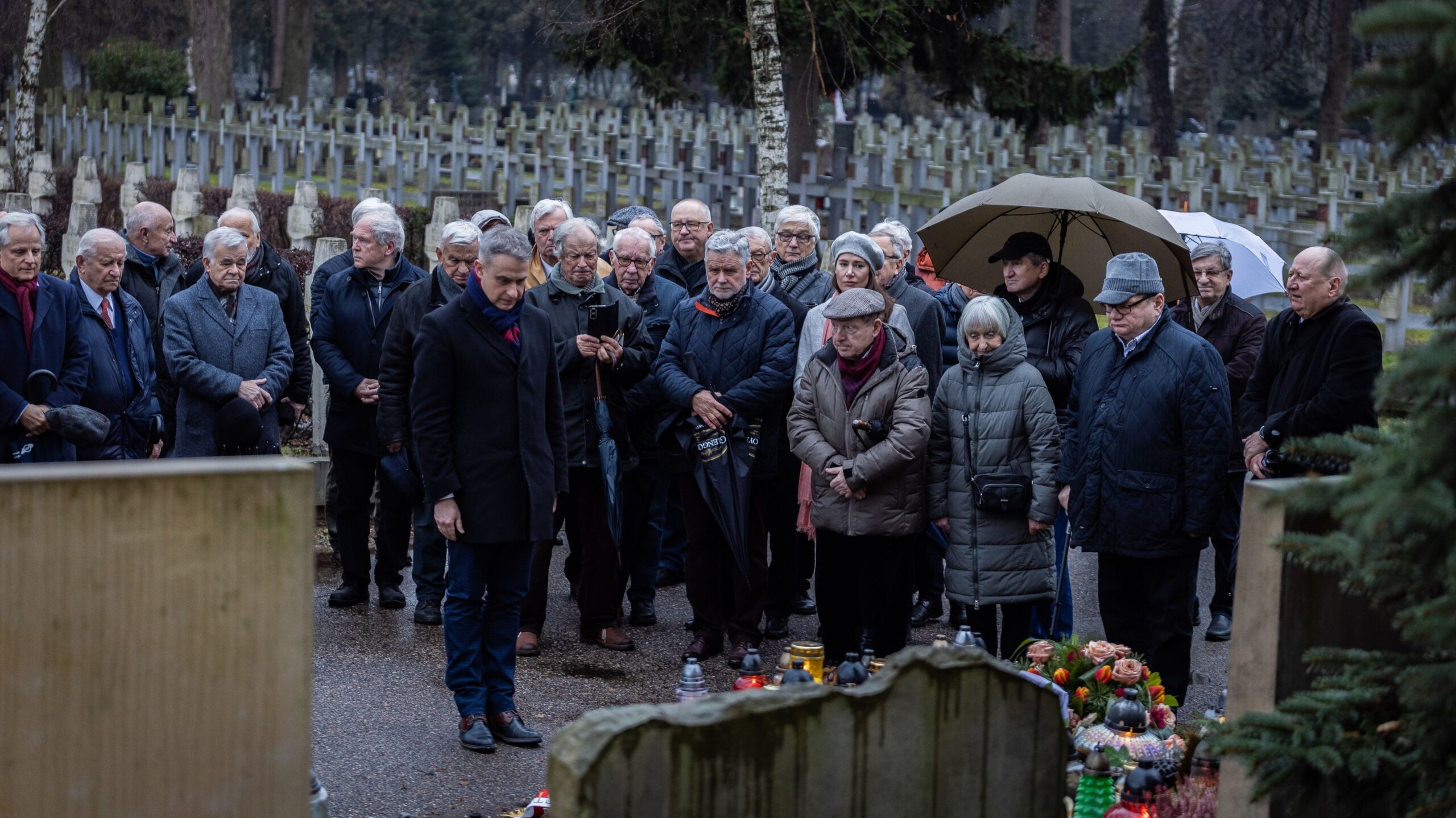 Na zdjęciu widoczna jest grupa ludzi zgromadzona na cmentarzu, w jedności i skupieniu, podczas ceremonii upamiętniającej zmarłych, z zapalonymi zniczami i parasolami w deszczową aurę.