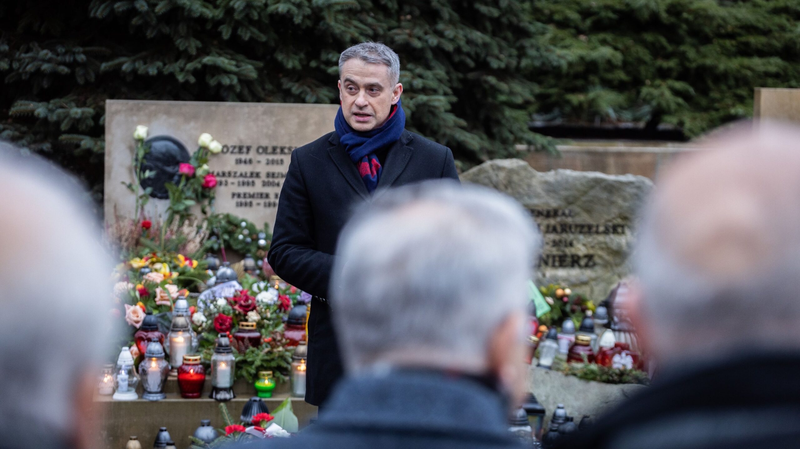 Mężczyzna w czarnym płaszczu i granatowym szaliku stoi przy kamiennym pomniku upamiętniającym, otoczony kwiatami i zapalonymi zniczami.