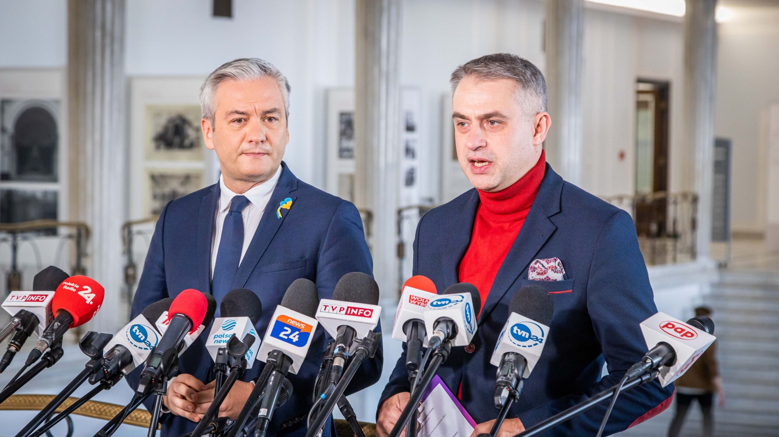 Na zdjęciu dwaj mężczyźni przemawiają na konferencji prasowej, otoczeni mikrofonami mediów w stylowym wnętrzu.