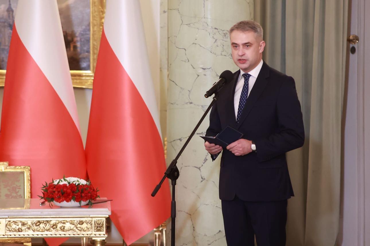 Mężczyzna w ciemnym garniturze przemawia do mikrofonu podczas zaprzysiężenia rządu, trzymając kartki, na tle stoją dwie flagi państwowe i ozdobna sala.