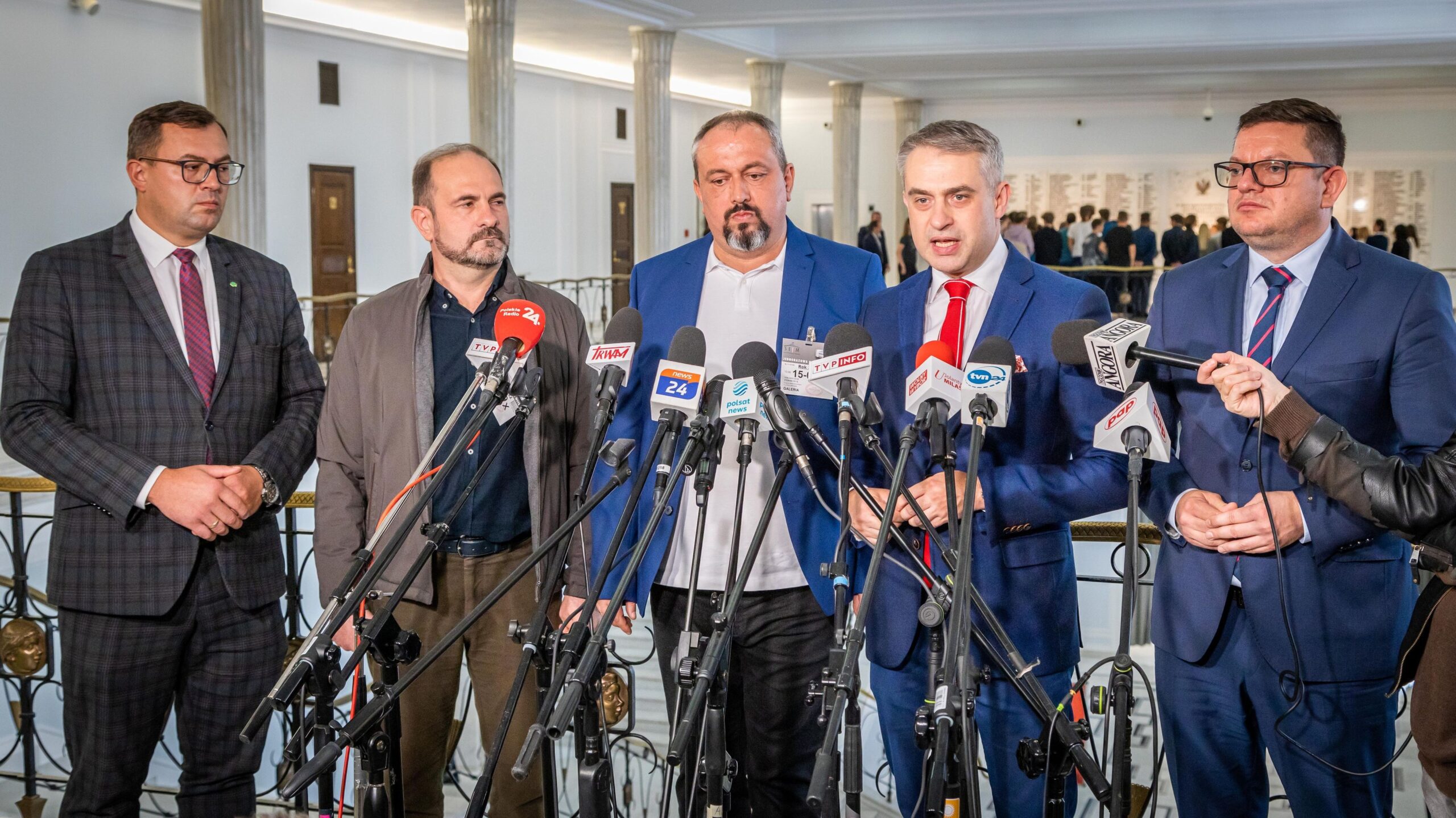 Czterej mężczyźni w garniturach stoją wśród mikrofonów podczas konferencji prasowej w eleganckim pomieszczeniu.