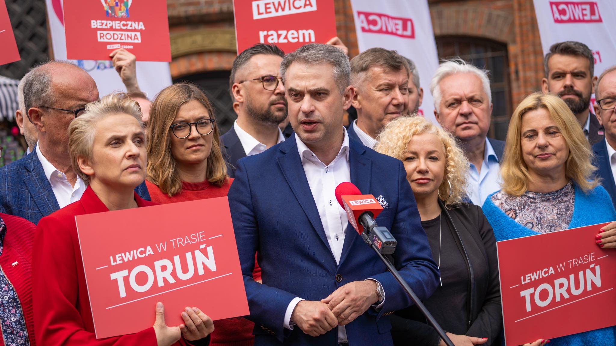 Grupa działaczy Lewicy w Kujawsko-Pomorskiem podczas konferencji prasowej, otoczona czerwonymi plakatami z napisem "LEWICA W TRASIE TORUŃ" i plakatem "BEZPIECZNA RODZINA", przemawia do mikrofonu.